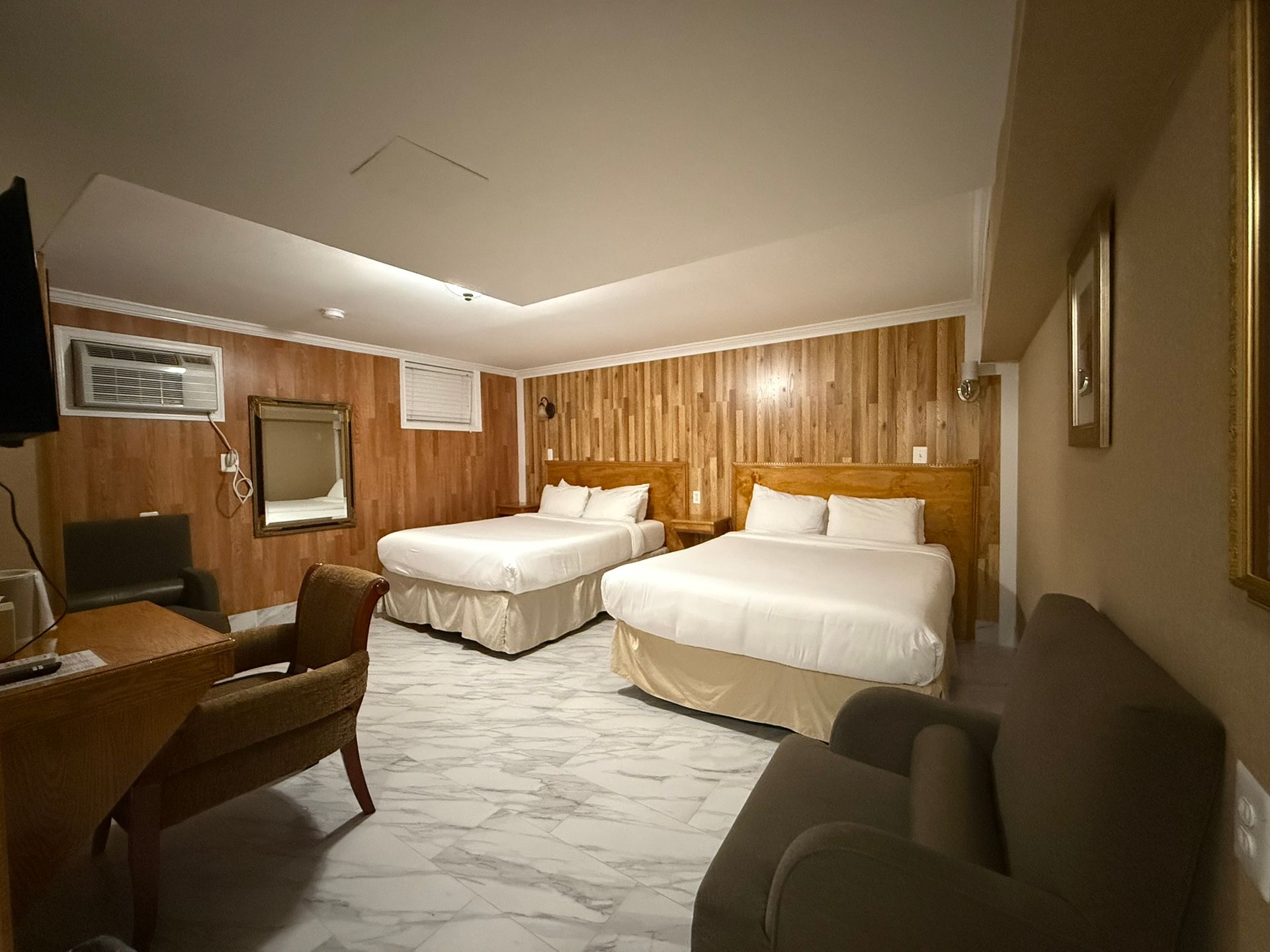 A hotel room with two double beds, a small sofa, and a desk. The walls are paneled with light wood and the floor is marble.