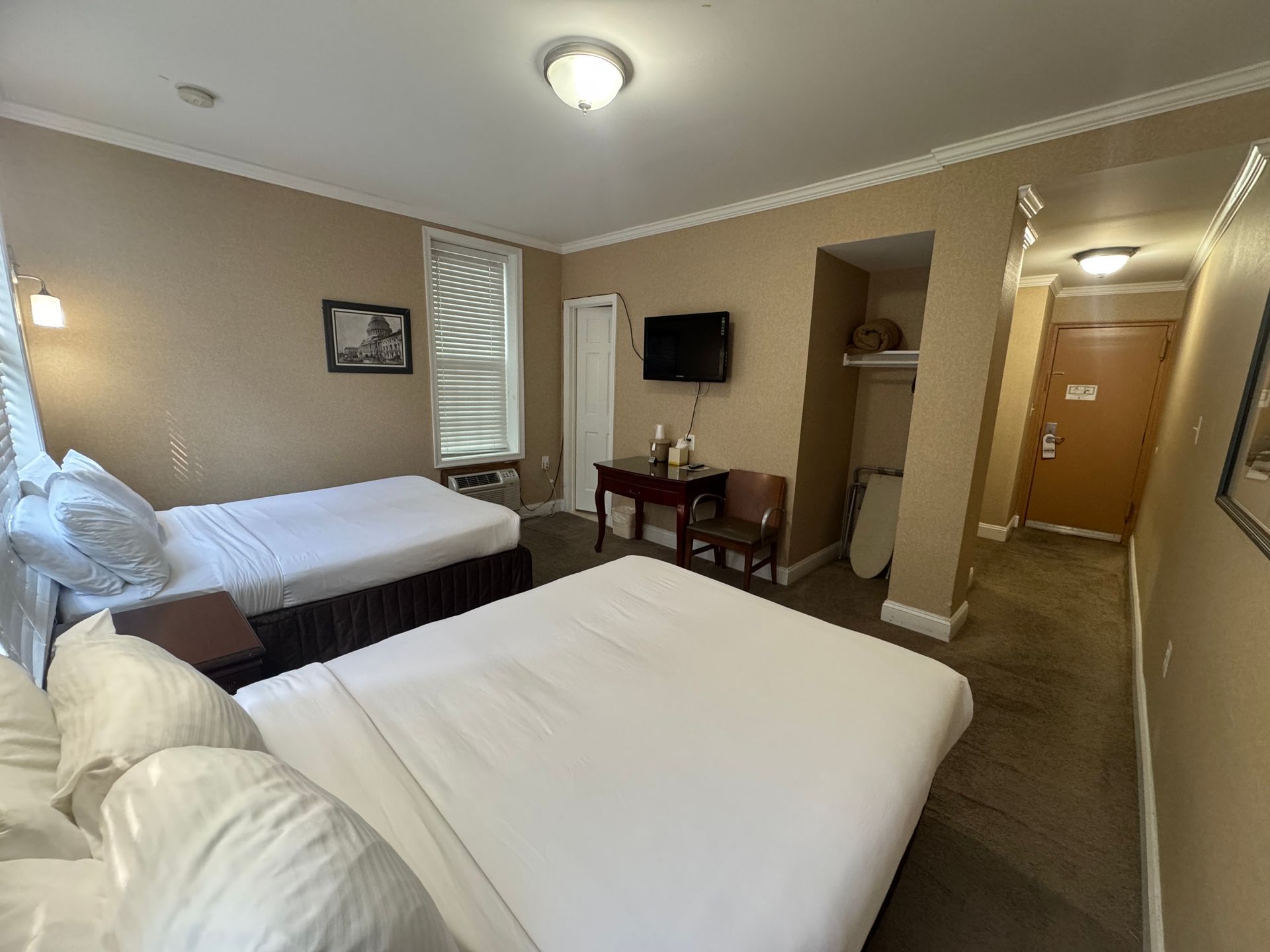 Hotel room with two beds, a small table, and a hallway leading to a door. Beige walls and white bedding.