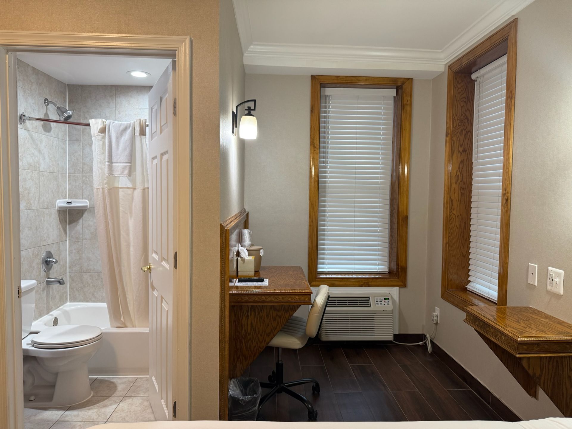Hotel room interior with a desk, windows, and an open bathroom door. A toilet and shower are visible in the bathroom.