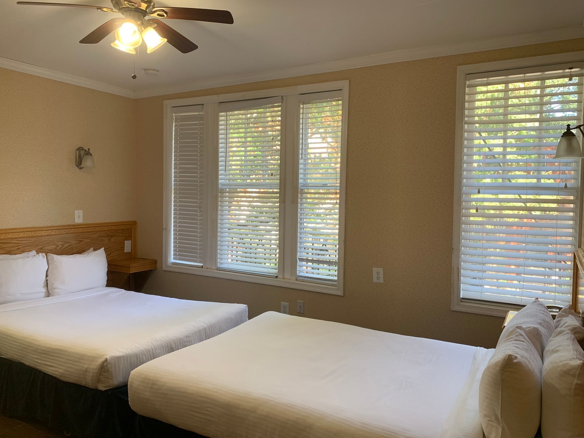 A hotel room with two queen beds, two windows with blinds, and a ceiling fan. The walls are beige.