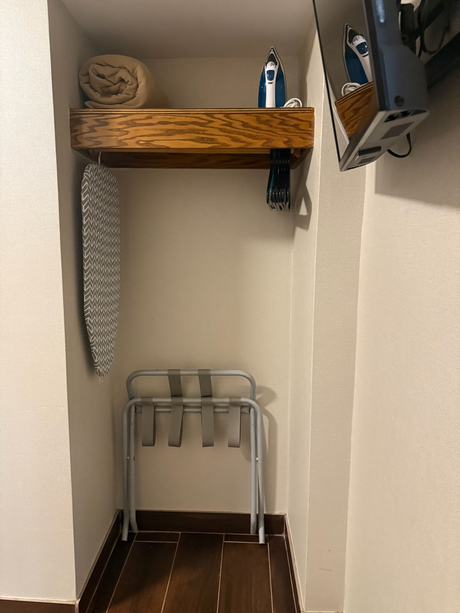 A hotel closet nook with a shelf holding a rolled towel, iron, and hanging cords. An ironing board hangs below.