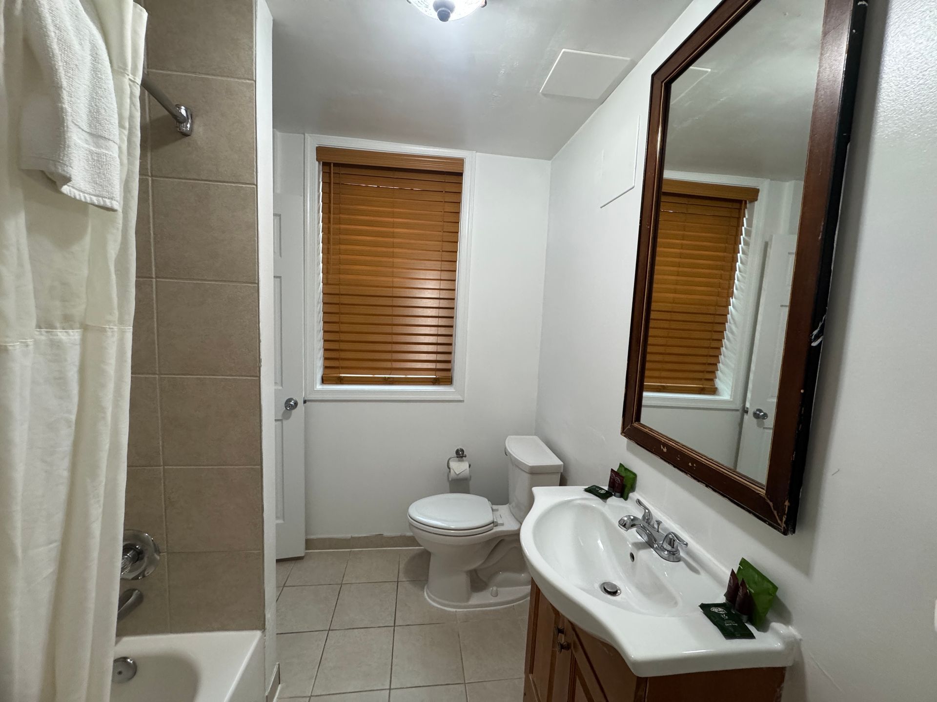 A bathroom with a vanity, toilet, and shower. The room has a wooden window shade and a large mirror.