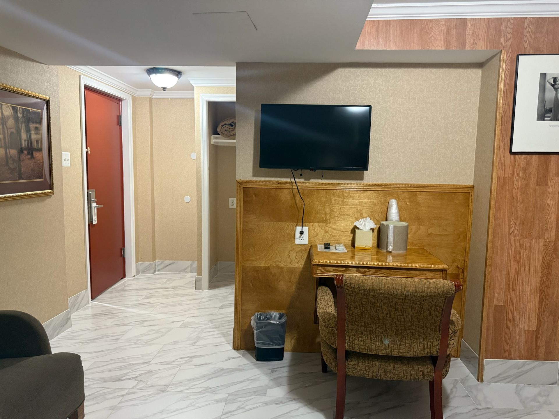 A hotel room with a desk, chair, TV, and doorway to a hall. The floor is tiled, and the walls are partially wood-paneled.