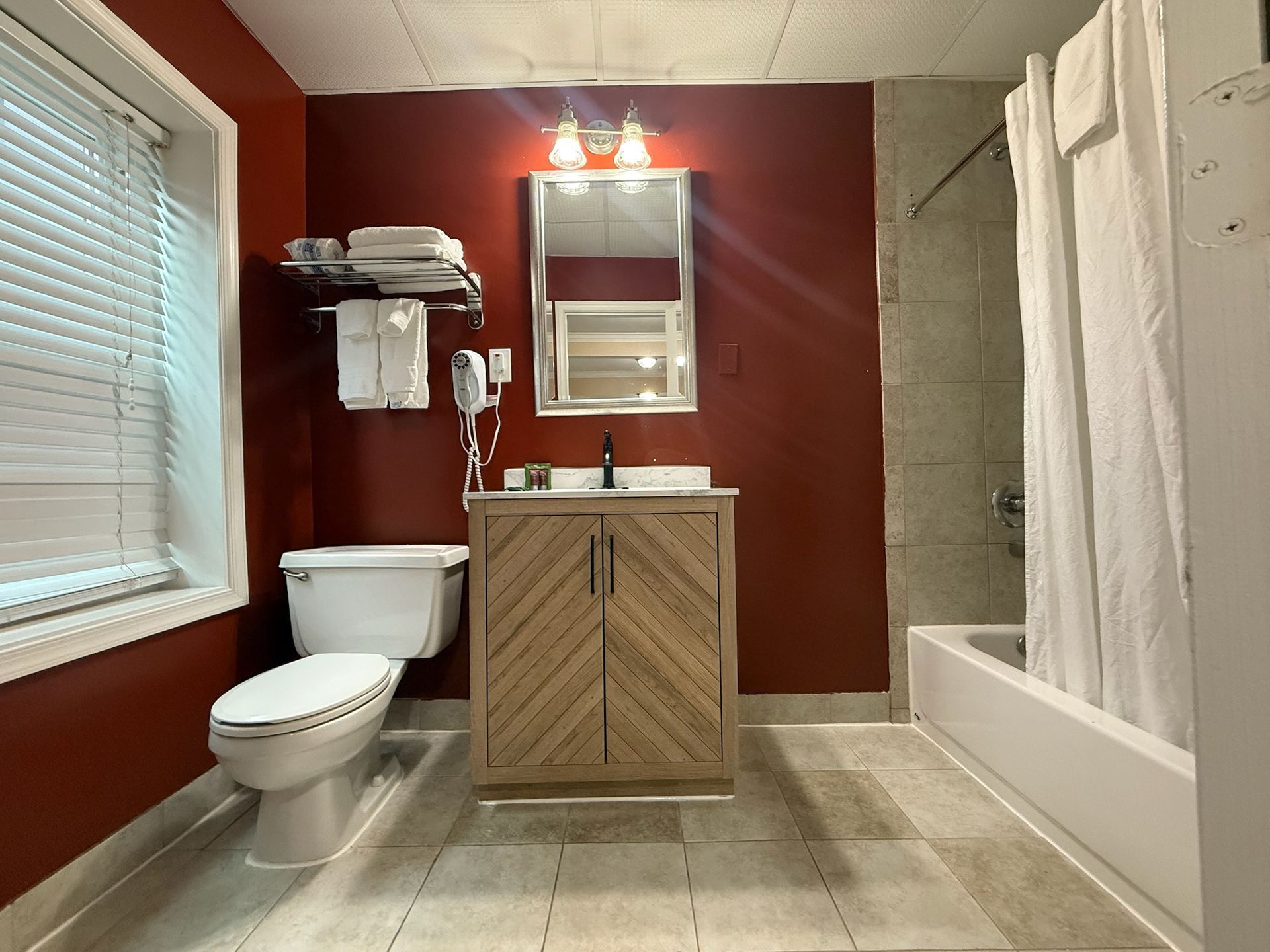 Bathroom with red walls, a light wood vanity, toilet, and bathtub. The room has a window with blinds.