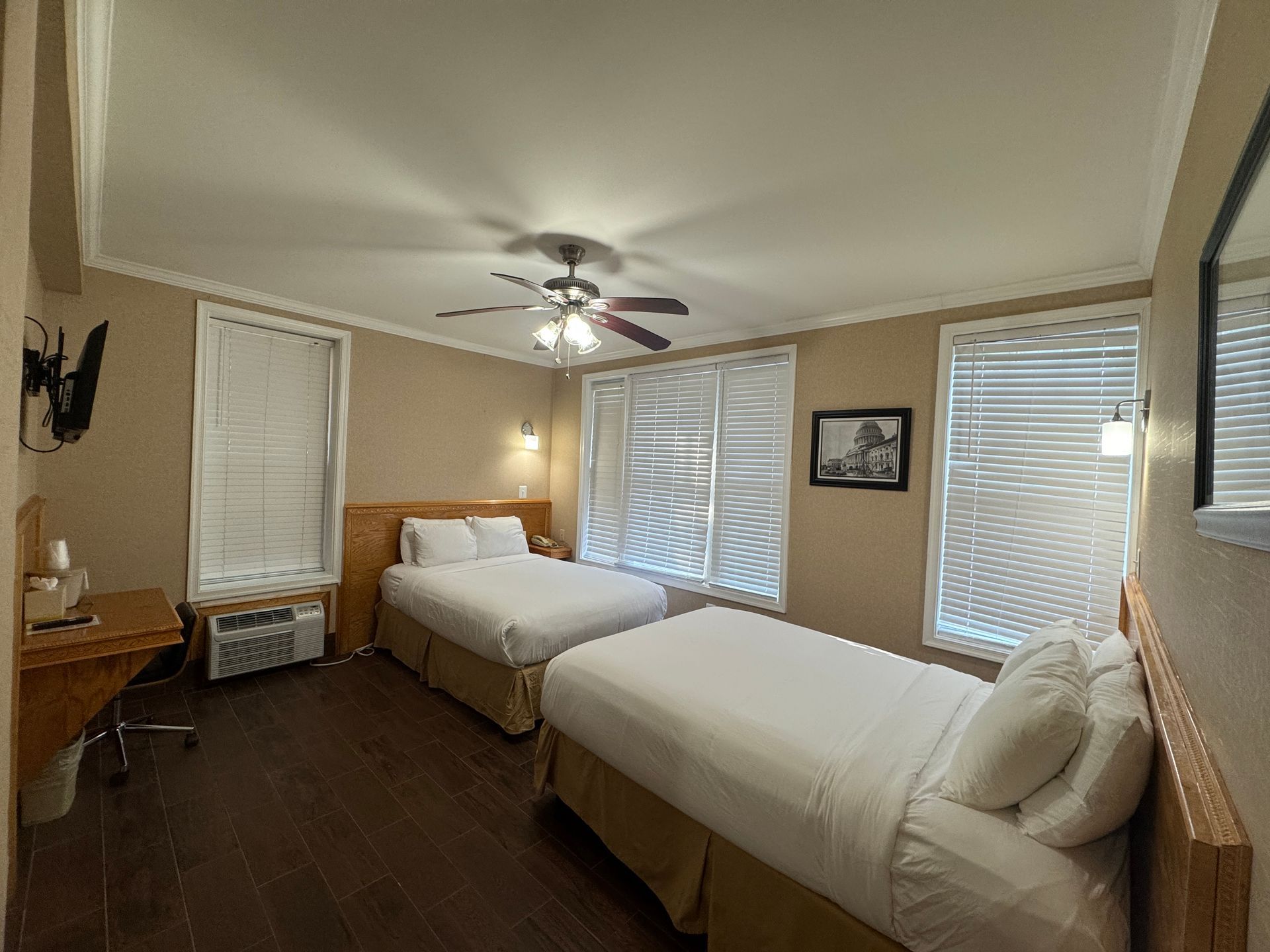 A hotel room with two beds, a desk, and blinds. The room has light brown walls, dark wood floors, and a ceiling fan.