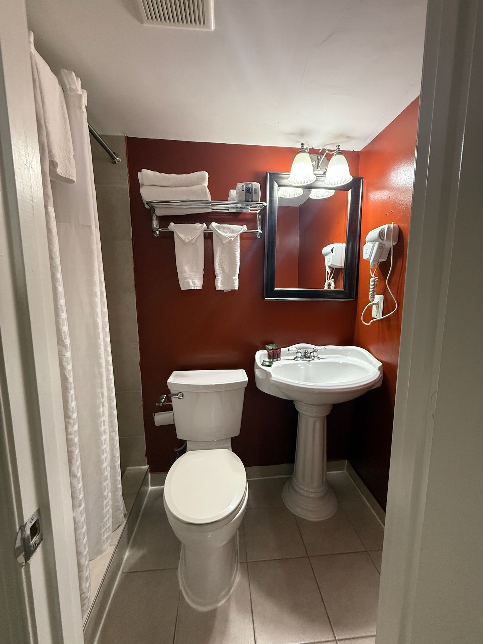 Bathroom with a toilet, pedestal sink, and towels on a rack, all against a reddish-brown wall.