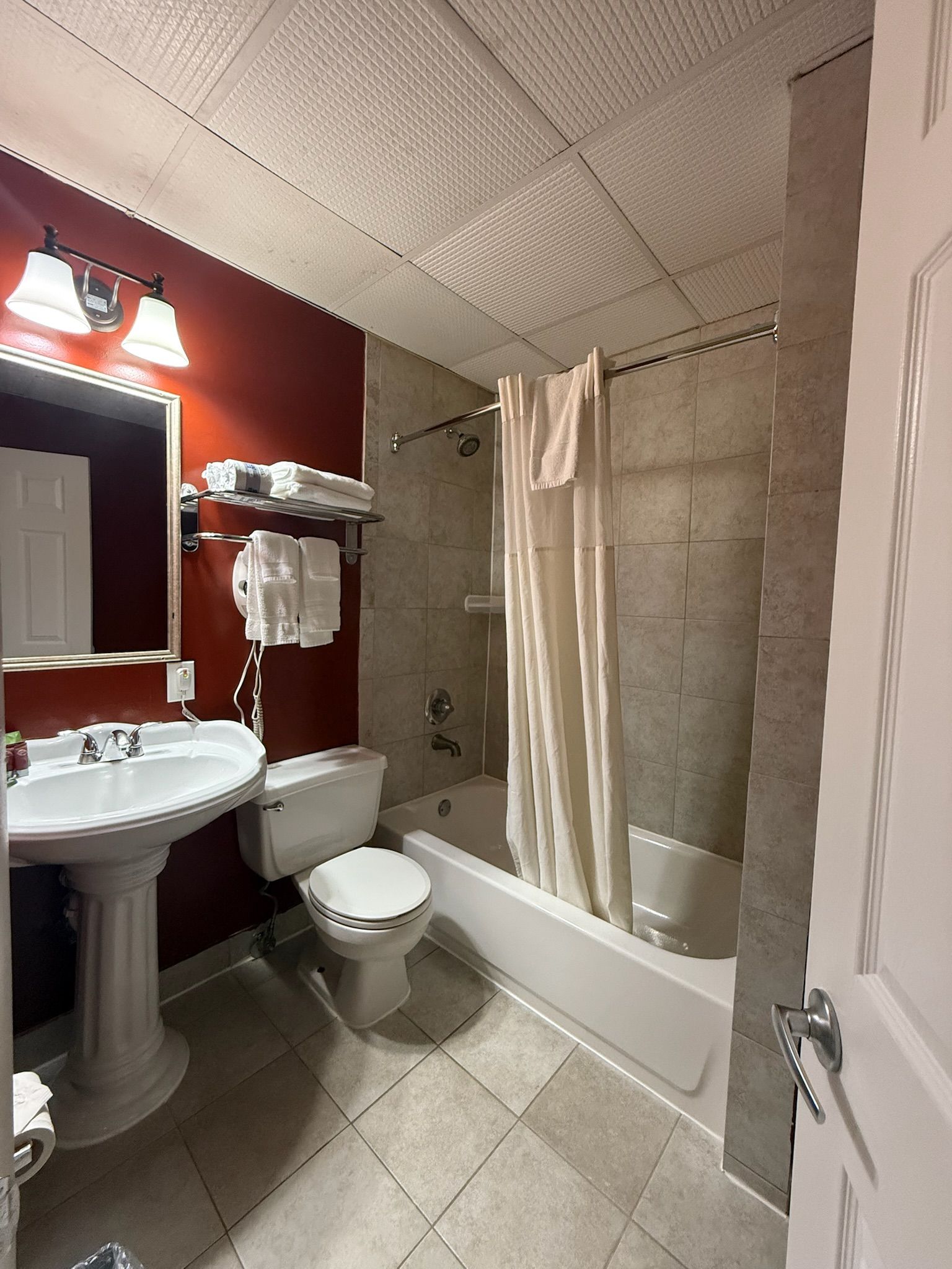 Small hotel bathroom with a pedestal sink, toilet, and shower/tub combo. Red accent wall, neutral tiles, and a white ceiling.