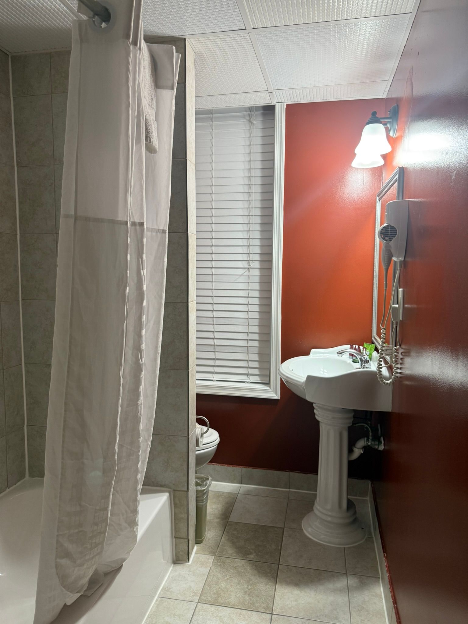 Bathroom with a white pedestal sink, orange walls, and a shower with a white curtain. A window with blinds is visible.