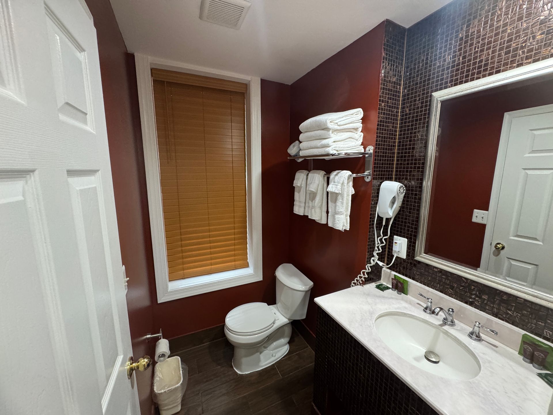 Bathroom with a toilet, sink, mirror, and a window with blinds. Dark red walls and light-colored towels are present.