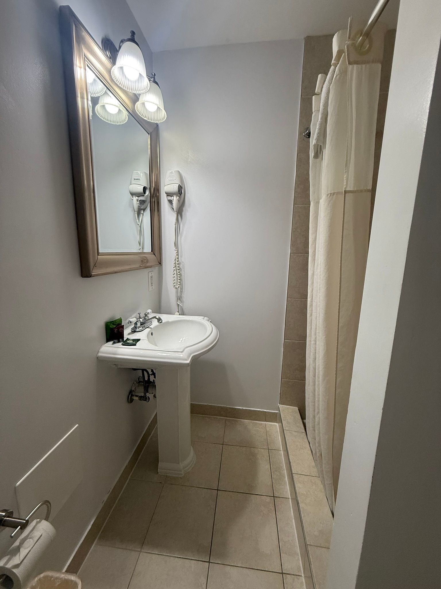 A small bathroom with a pedestal sink, mirror, and shower. The walls are light gray, and the floor is tiled.