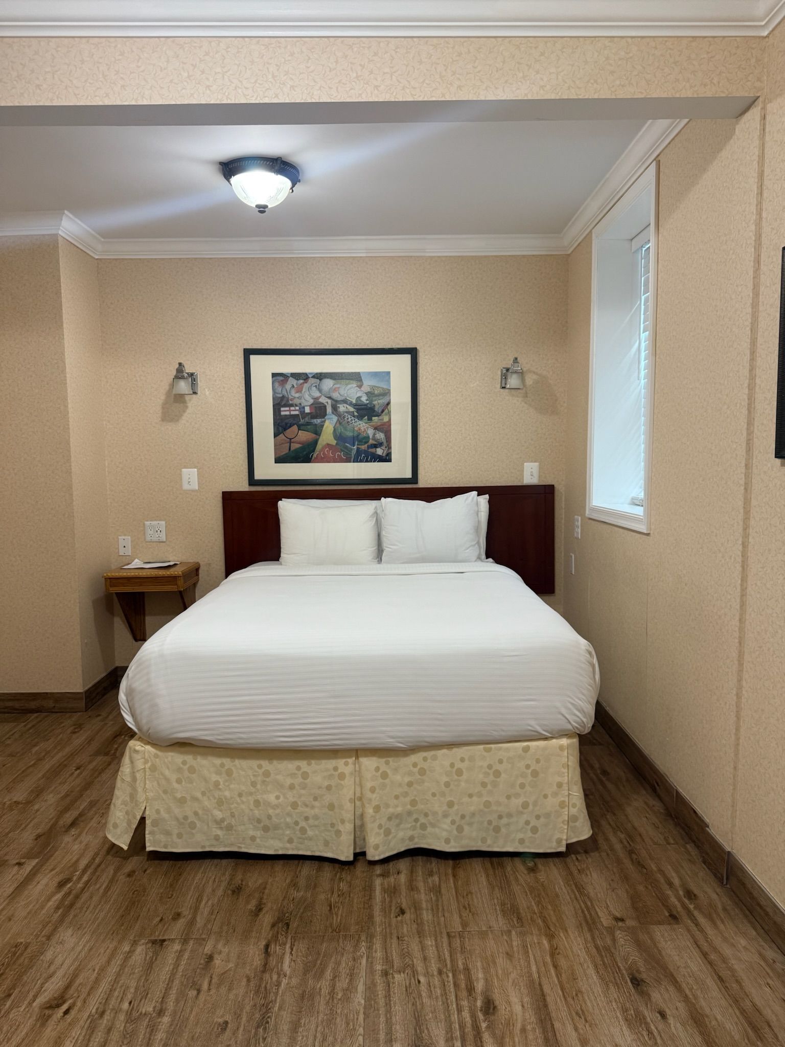 A hotel room with a bed, nightstand, and framed art. The walls are patterned, and the floor is wood-like.