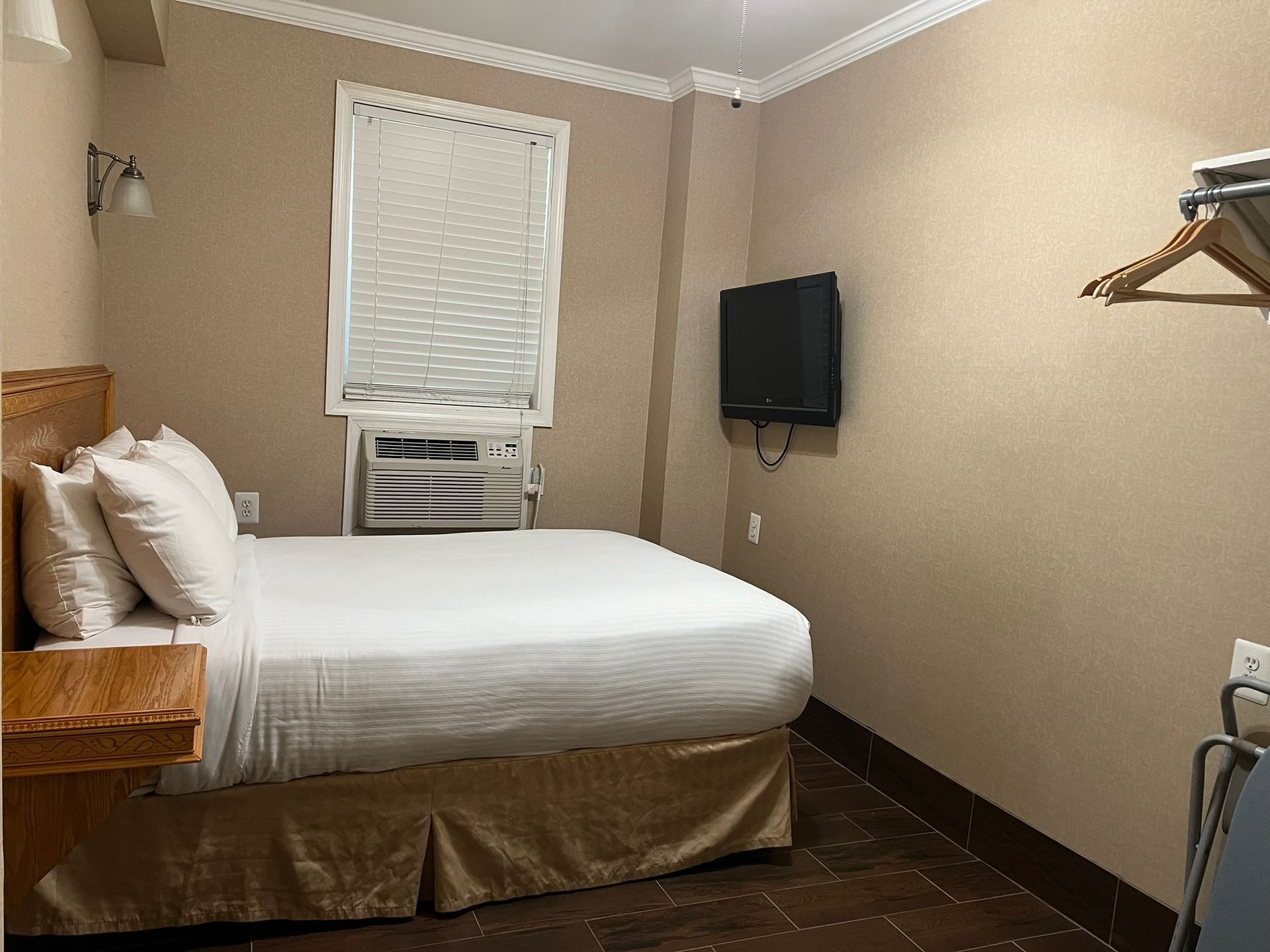 A hotel room with a bed, television, and window with blinds. The walls are beige, and the bed has white bedding.