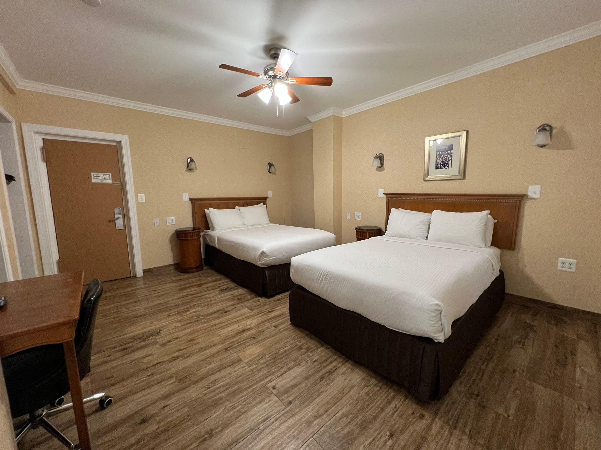 A hotel room with two beds, a desk, and a closed door. The walls are beige, and the floor is wood-like.