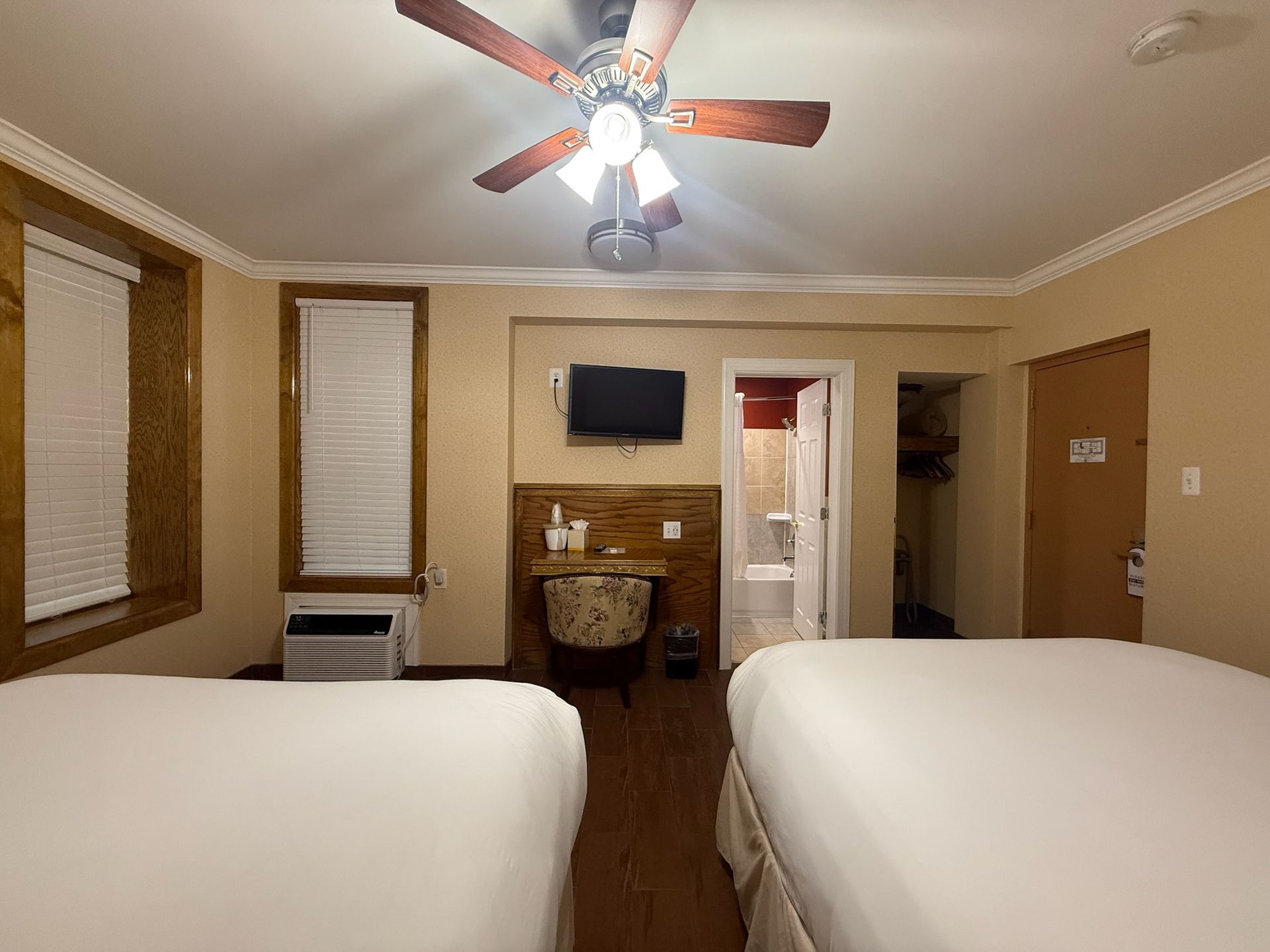 A hotel room with two beds, a desk, and a ceiling fan. The bathroom door is open.