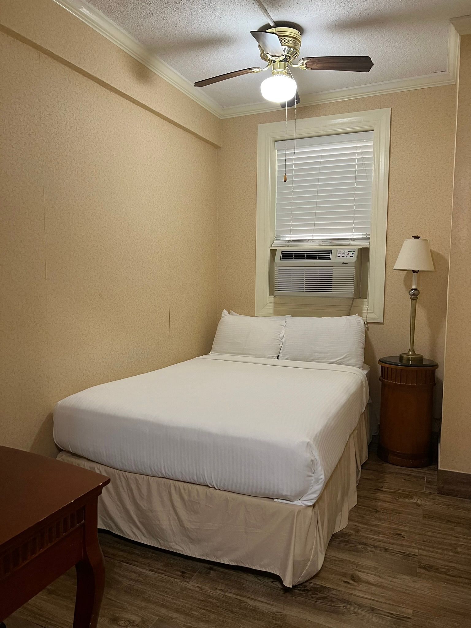 A small hotel room with a bed, window AC unit, and a lamp on a bedside table. Beige walls and a ceiling fan are visible.