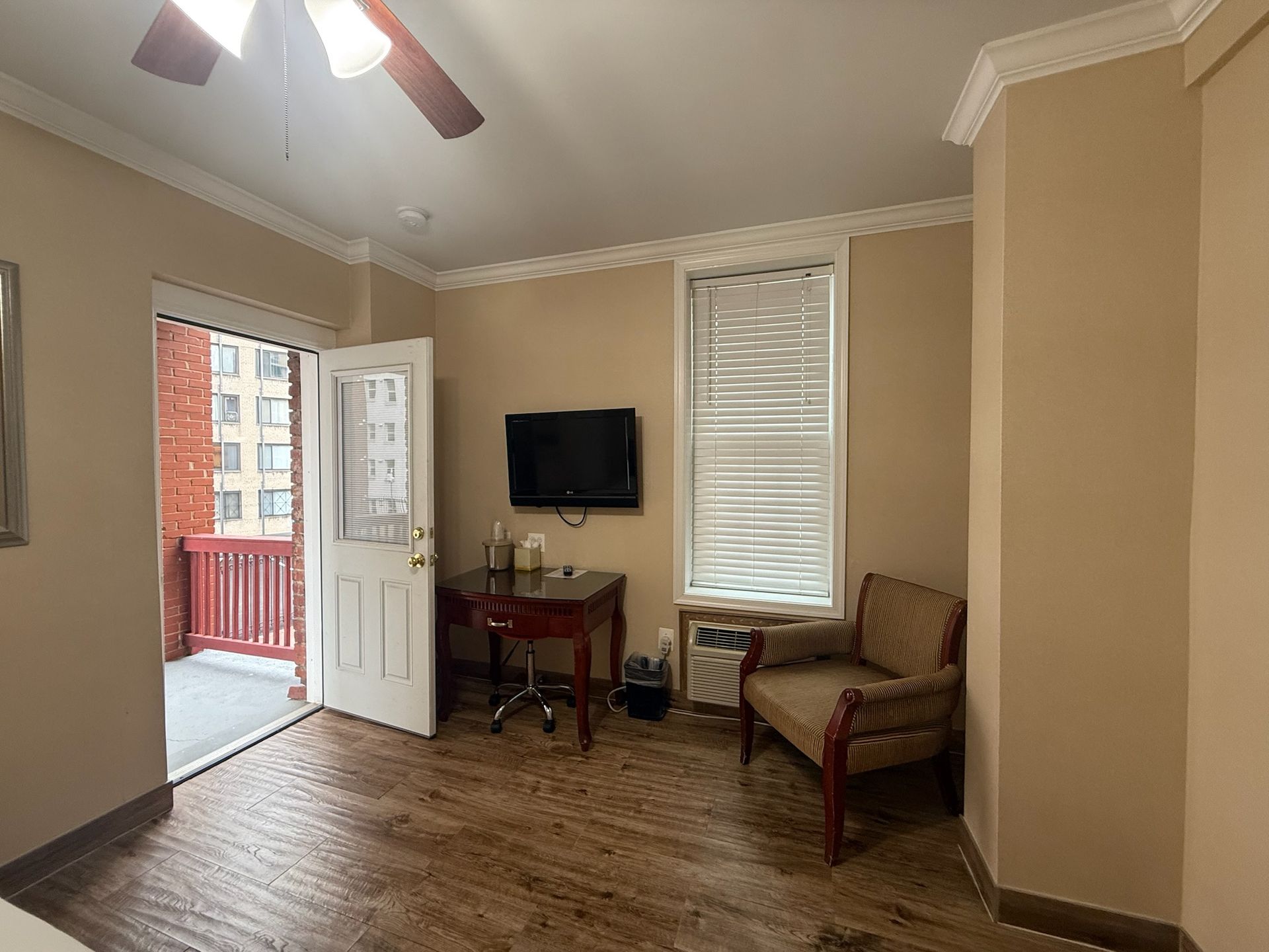 A hotel room with a balcony. The room has a desk, chair, and TV. The walls are beige, and the floor is wood.