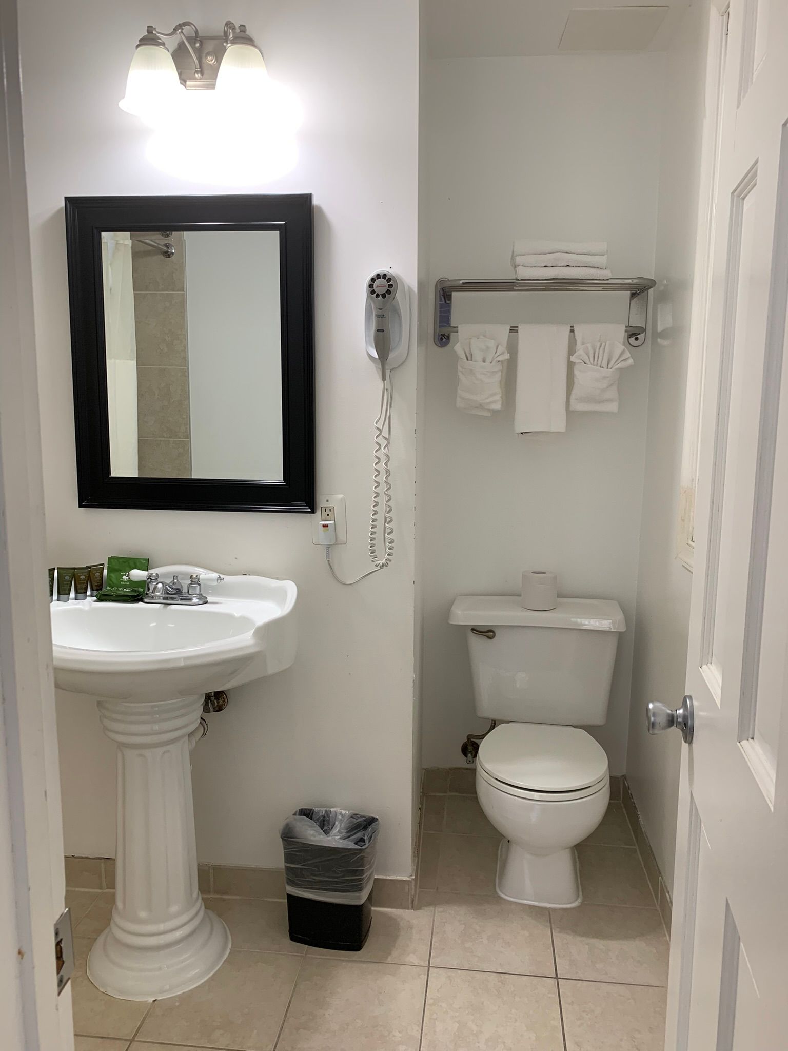 Bathroom with pedestal sink, mirror, toilet, and towel rack. Walls are white, floors tan tile. 