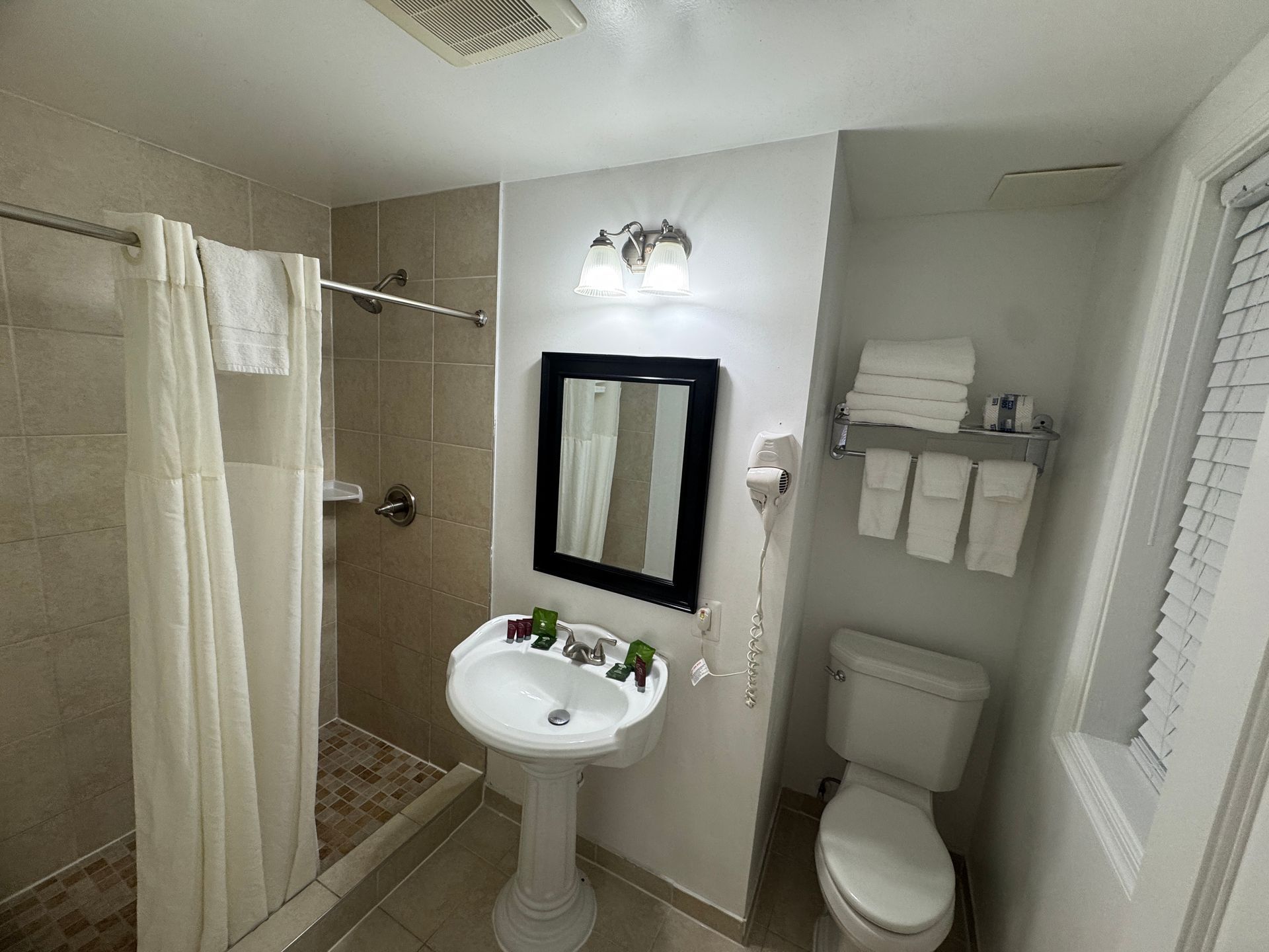 A small bathroom featuring a pedestal sink, toilet, shower with a curtain, and a window. White and tan color scheme.