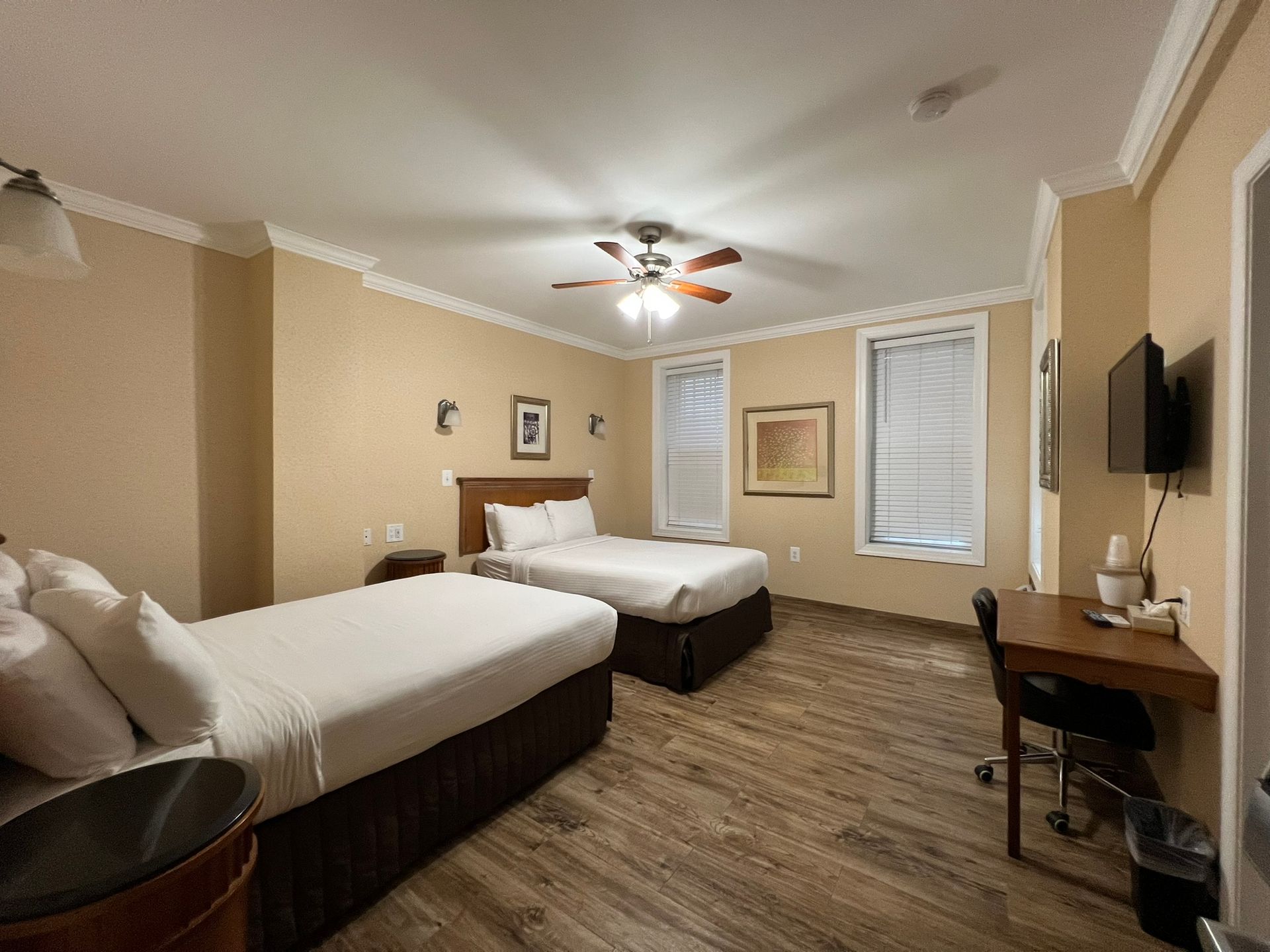 Hotel room with two beds, a desk, and a television. The room has tan walls, crown molding, and wood-look flooring.