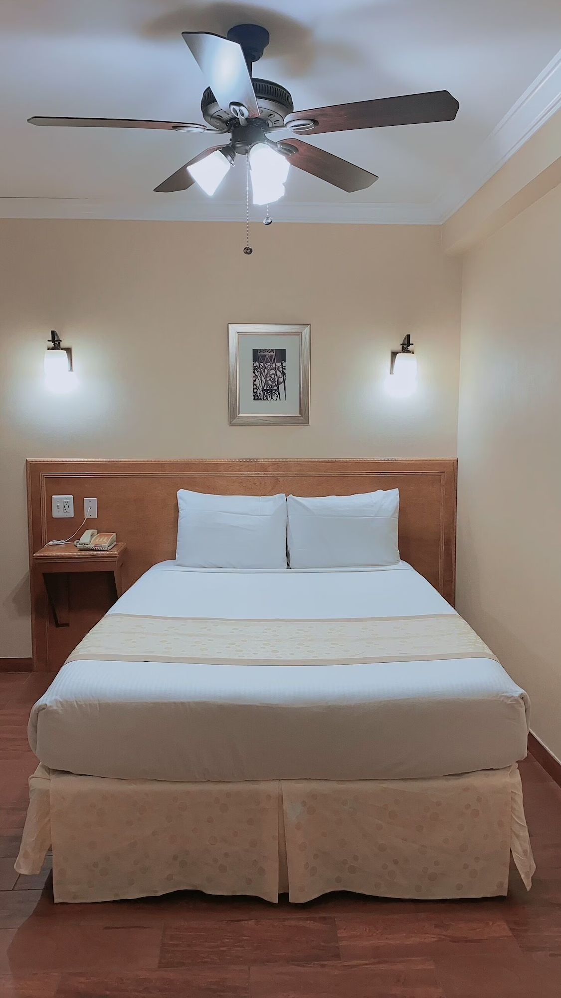 Hotel room with a bed, nightstand, artwork, and a ceiling fan.  The room is lit by wall-mounted lamps and has a wooden floor.