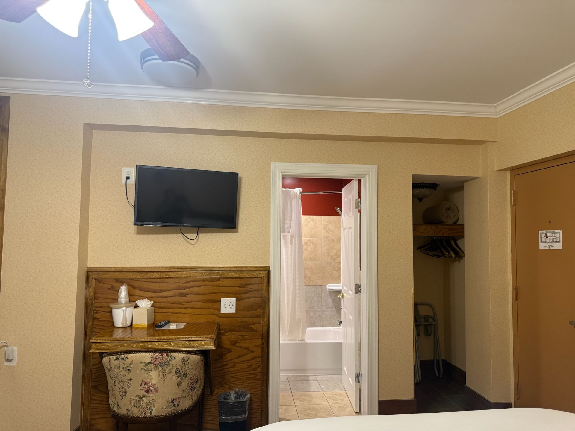 Hotel room interior. A small desk with a floral-patterned chair is under a mounted TV. The bathroom and closet are visible.