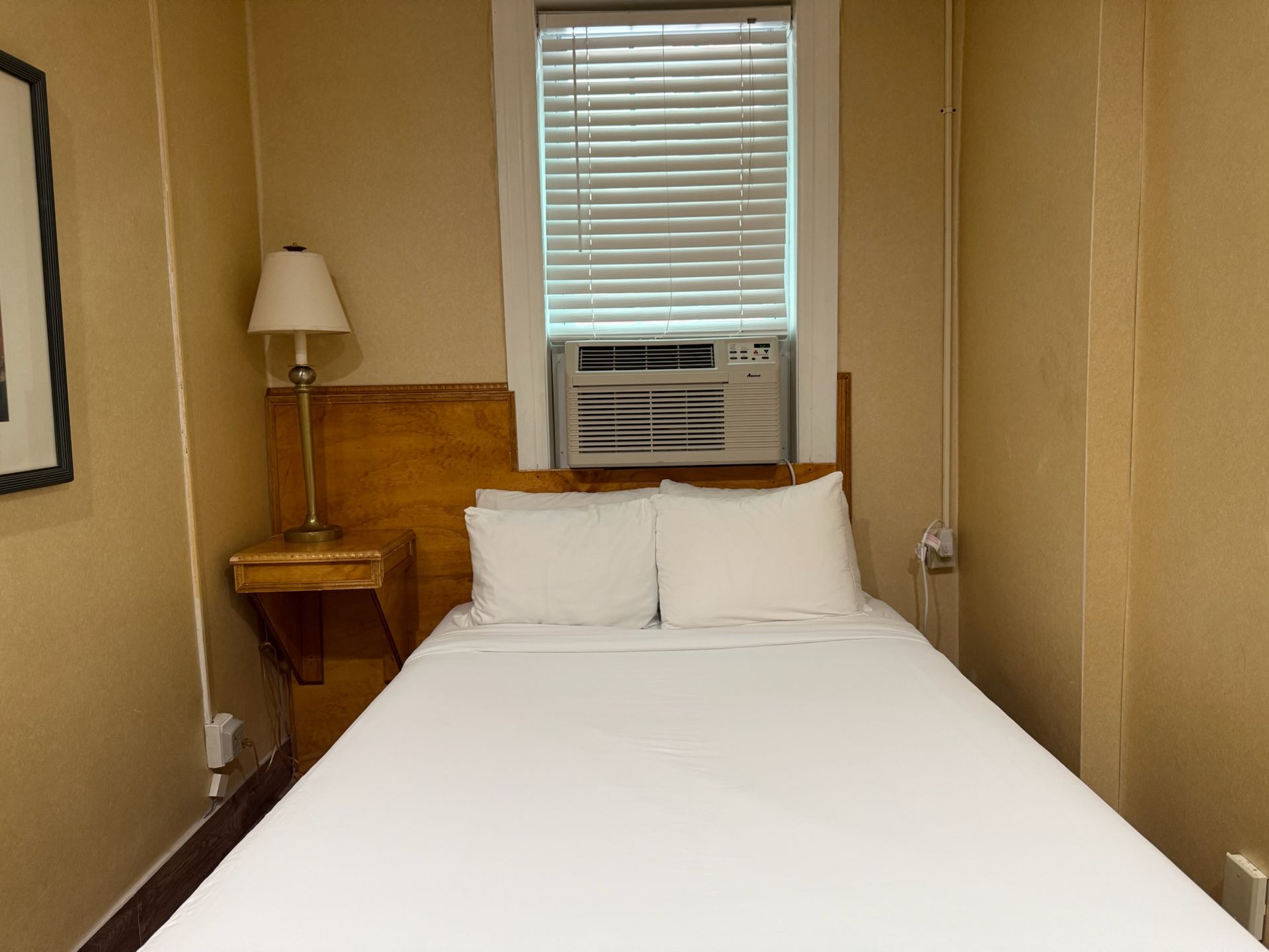 A small, simple hotel room with a bed, nightstand, and a window with an AC unit. Pale yellow walls.