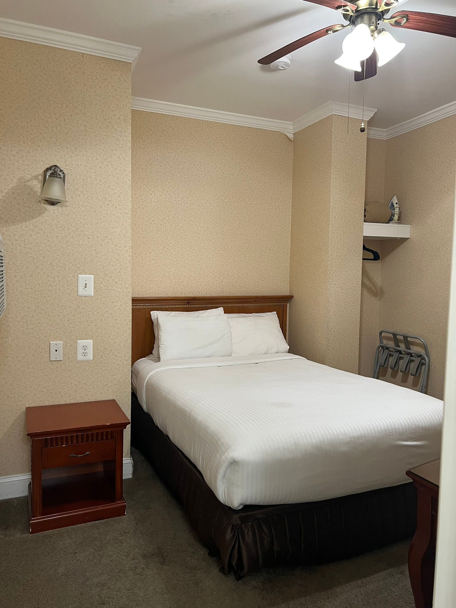 A small hotel room with a double bed, nightstand, and ceiling fan. Beige walls, brown bed skirt and furniture.