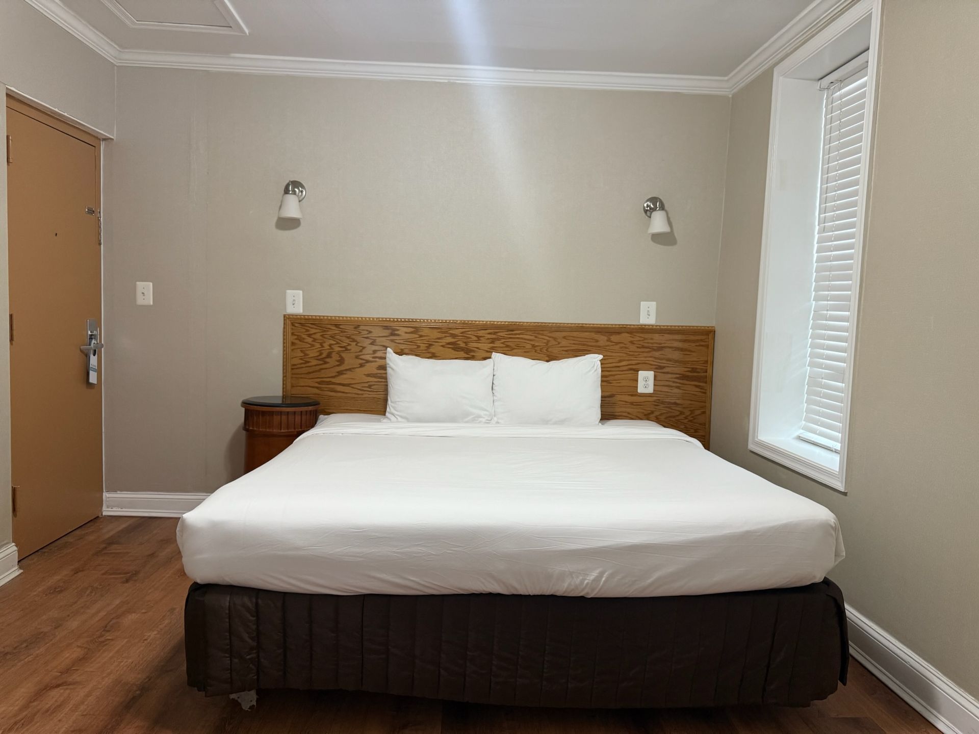 A hotel room featuring a made bed with white linens and wooden headboard. A nightstand is beside the bed.