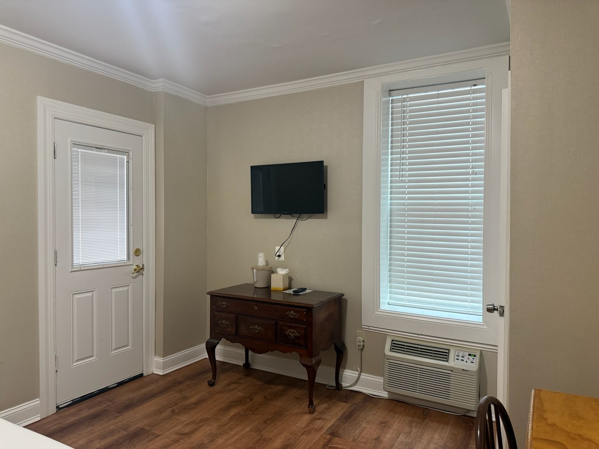 Small room with a TV, antique dresser, window with blinds, and a door with blinds; tan walls and wood-look flooring.