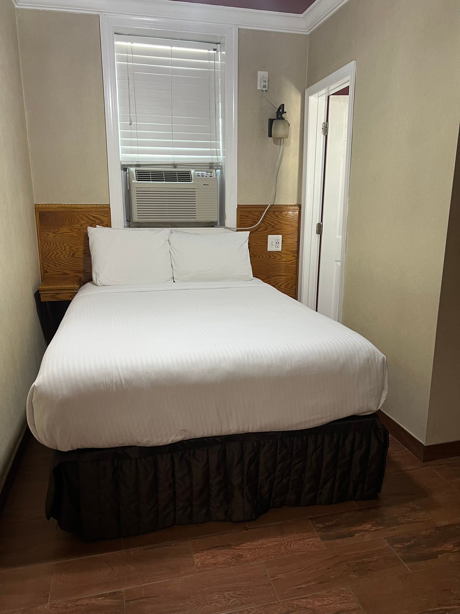 A small hotel room with a bed, window, and doorway. The bed has white linens and a brown bed skirt.