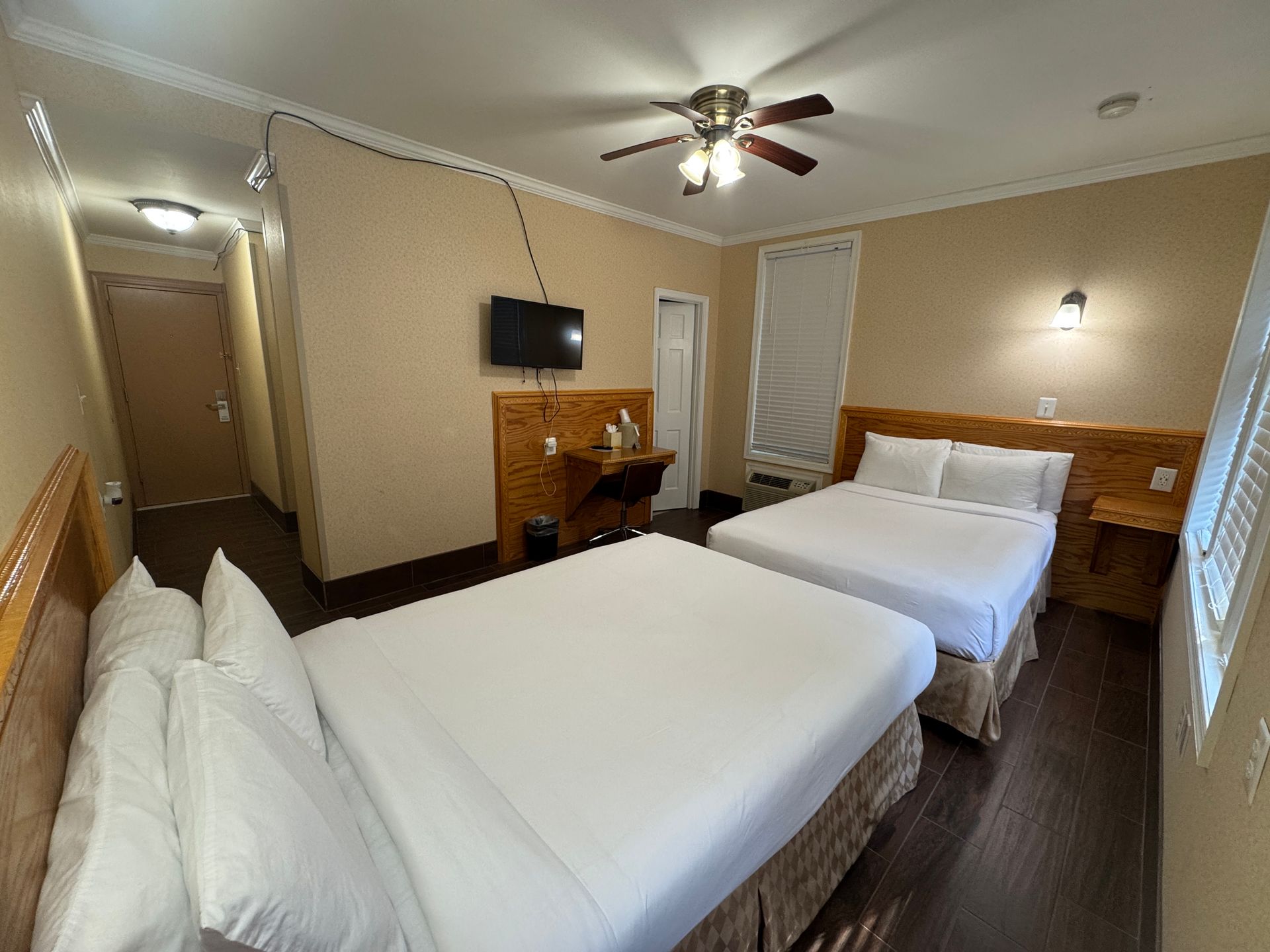 A hotel room with two double beds, a small desk, and a television. The walls are beige, and the floor is dark wood.