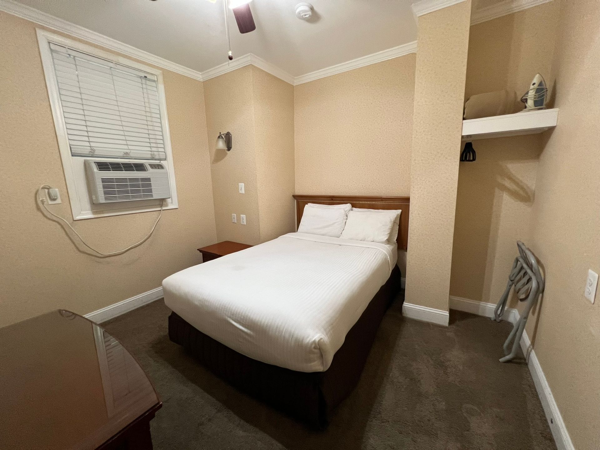 A small hotel room with a bed, window AC unit, and a dark carpet. The walls are a light beige color.