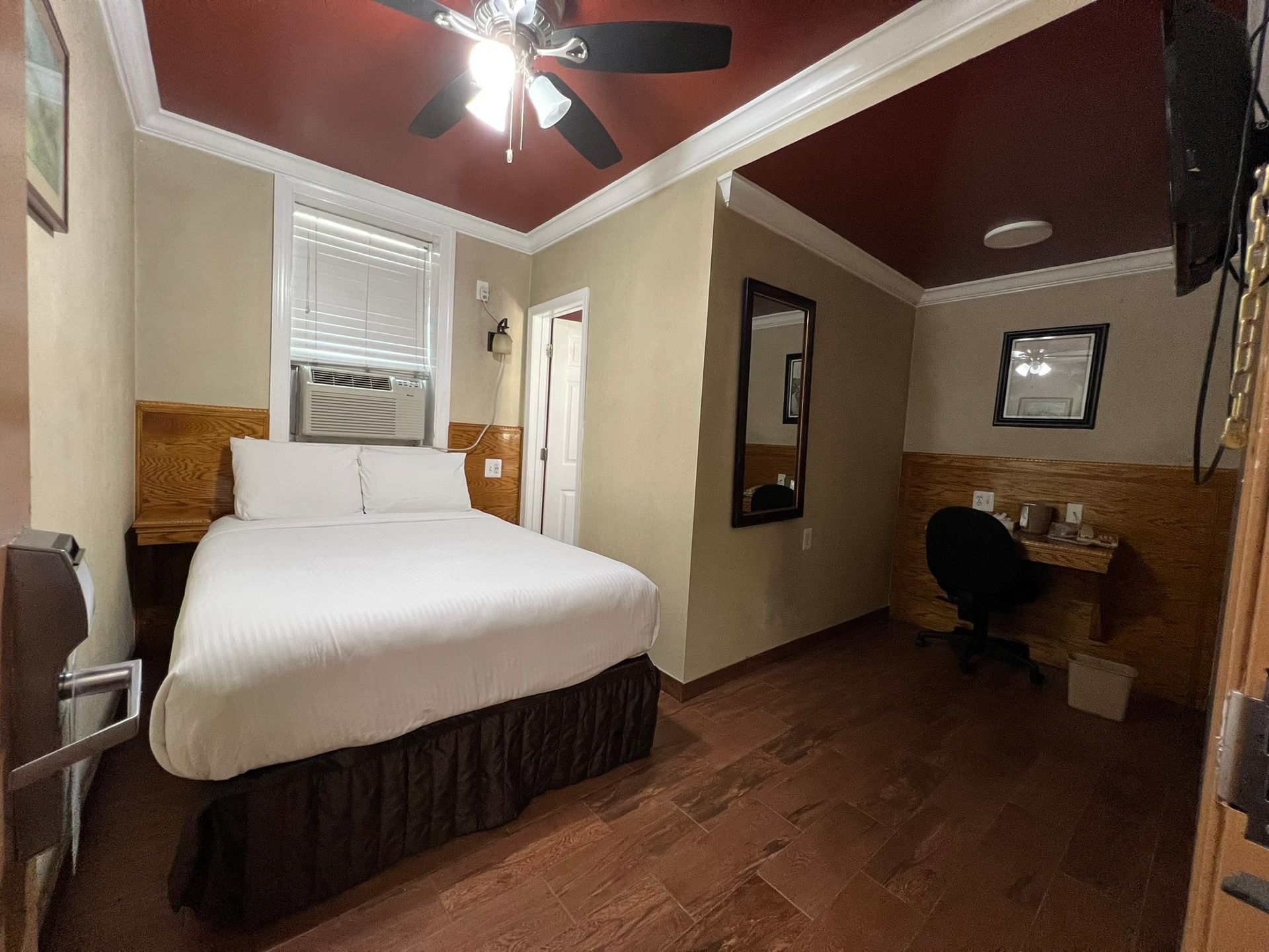 A hotel room with a bed, desk, mirror, and window. Red ceiling, brown floor, and beige walls.