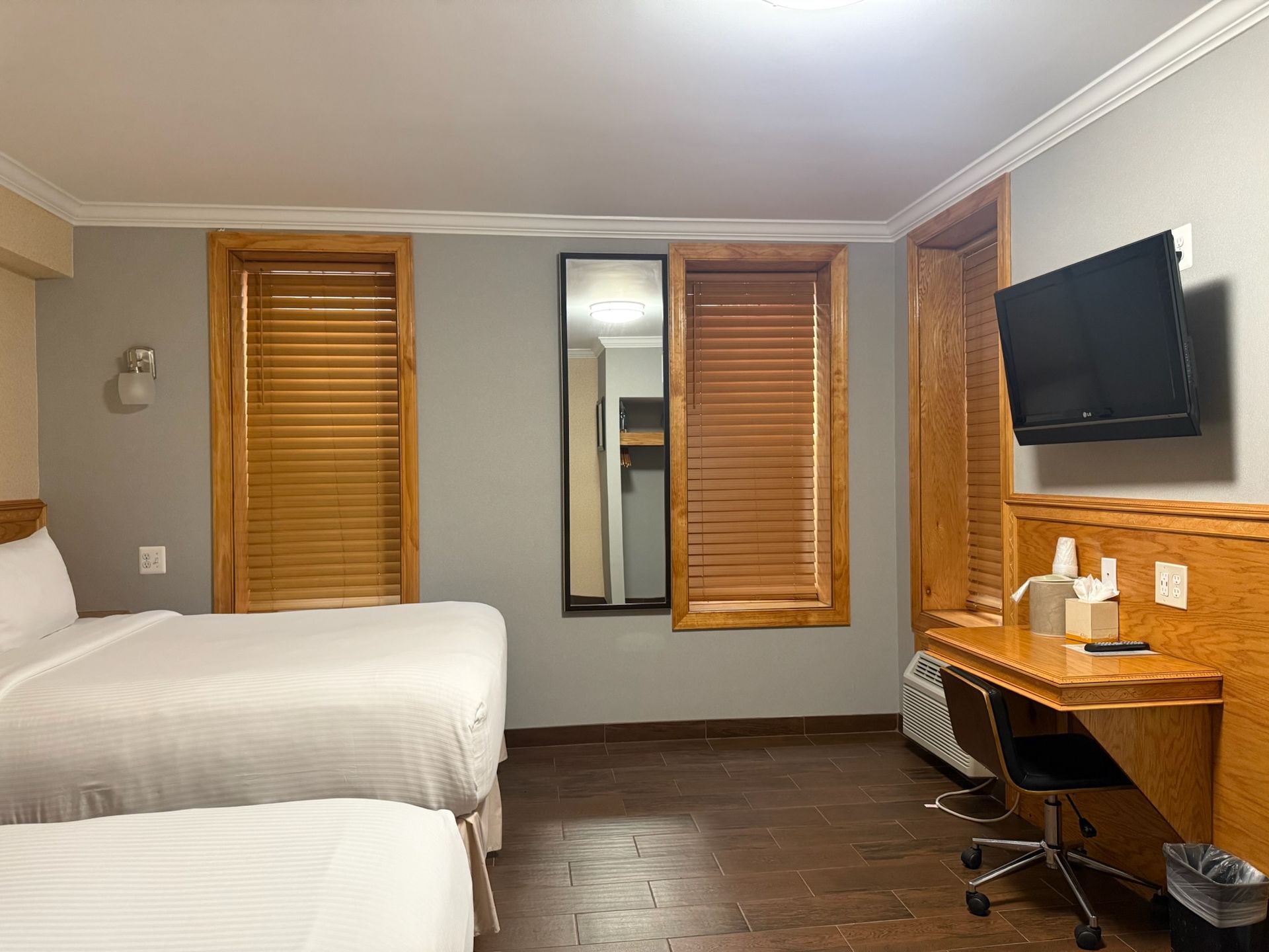 Hotel room with two beds, wooden blinds, and a desk with a mounted TV. The walls are gray with wood trim.