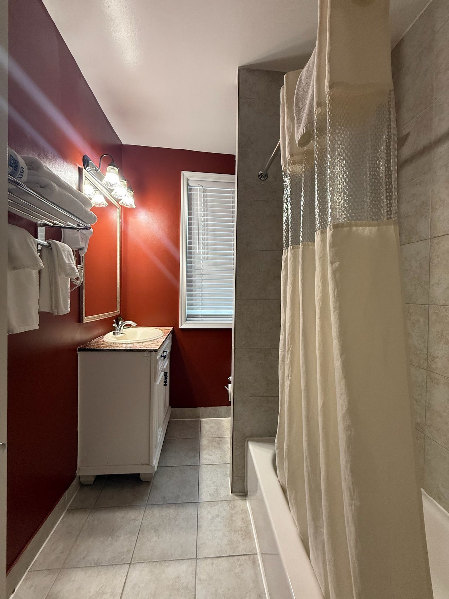 Bathroom with red walls, white vanity, and a shower with a beige curtain. A window with blinds is at the end of the room.