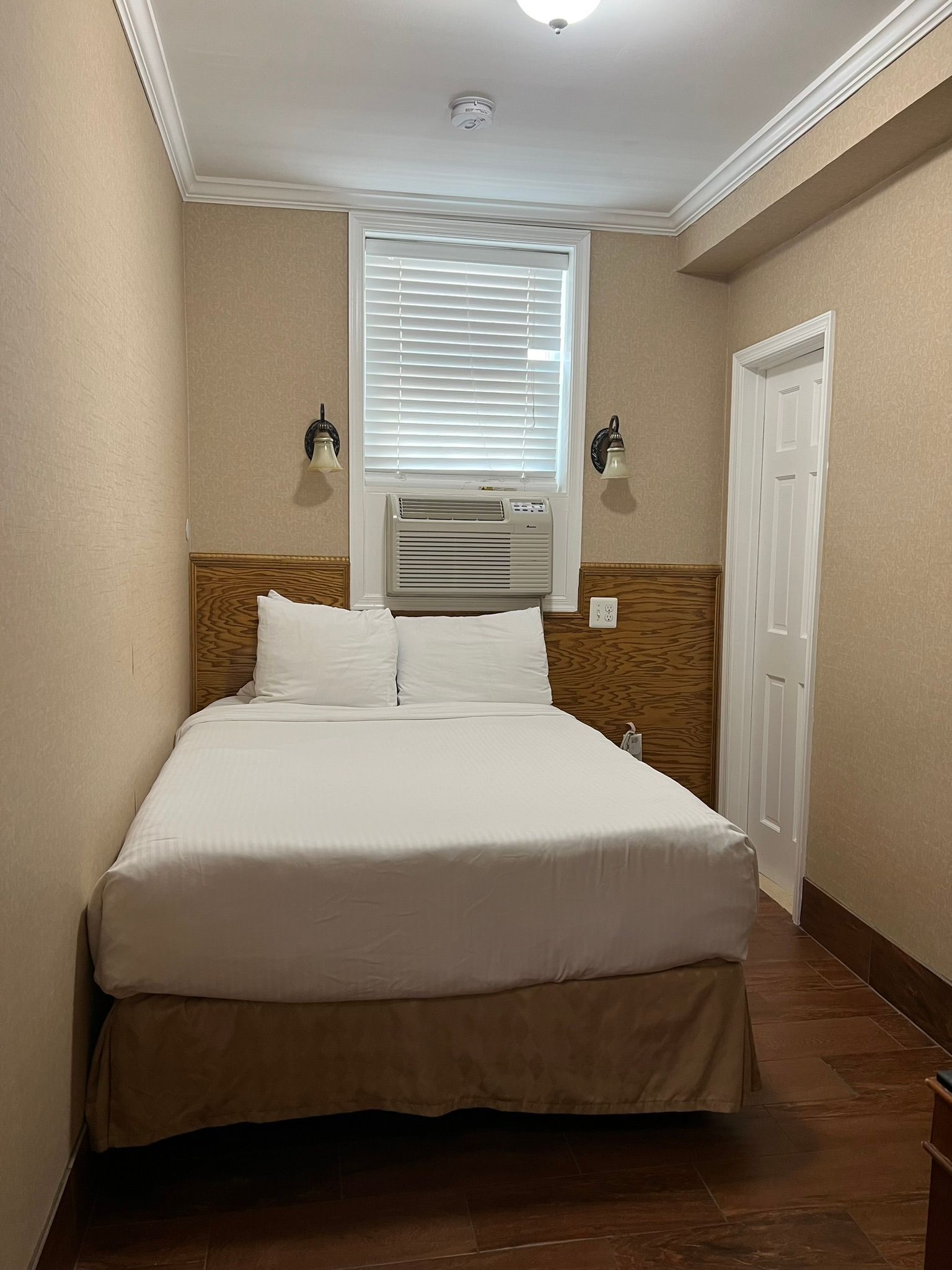 Small hotel room with a bed, window, and door. Beige walls, brown bed skirt, and a wooden headboard.