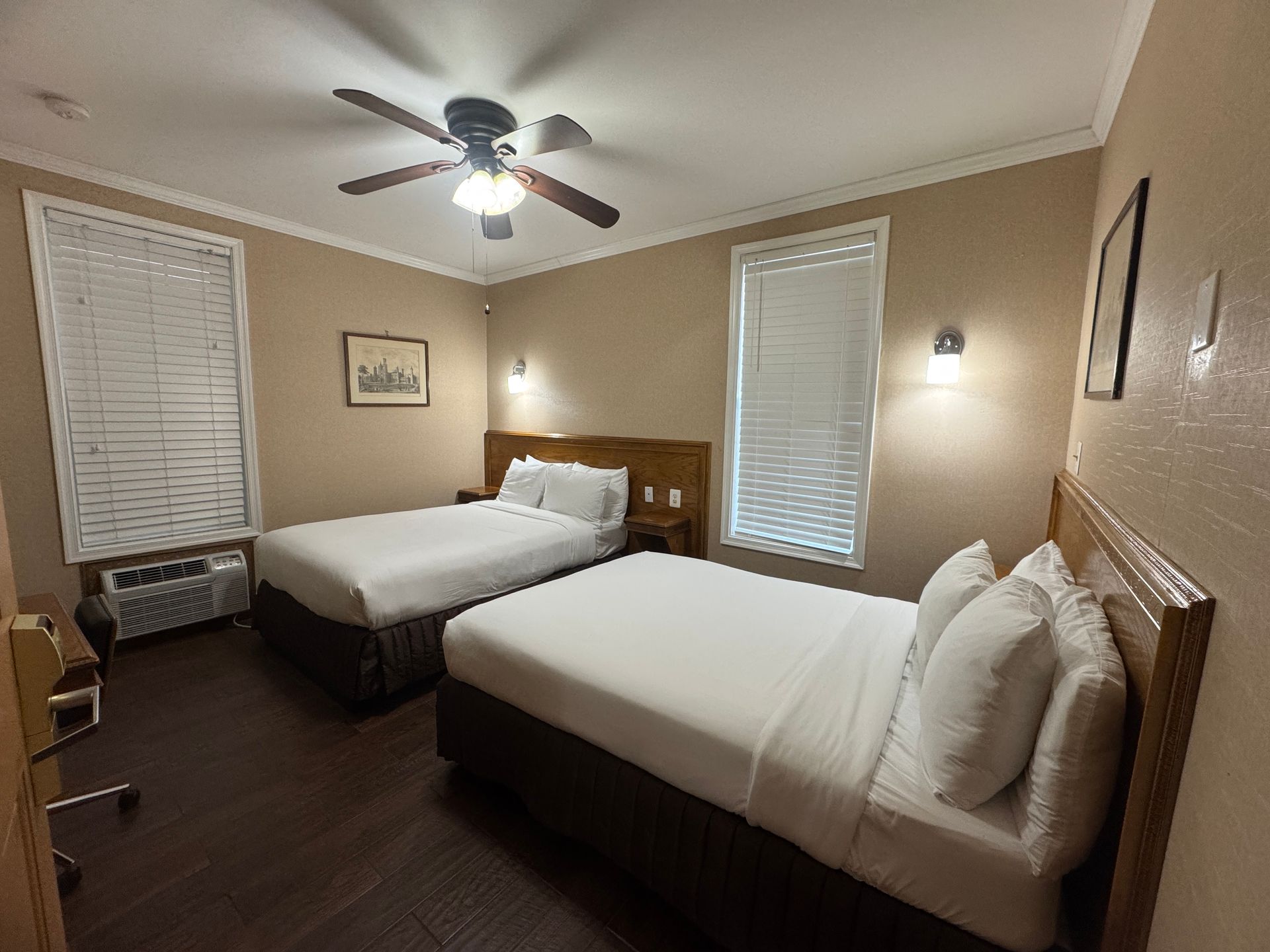 Hotel room with two double beds, brown wooden headboards, and dark wood floors. Beige walls, white blinds, and a ceiling fan.