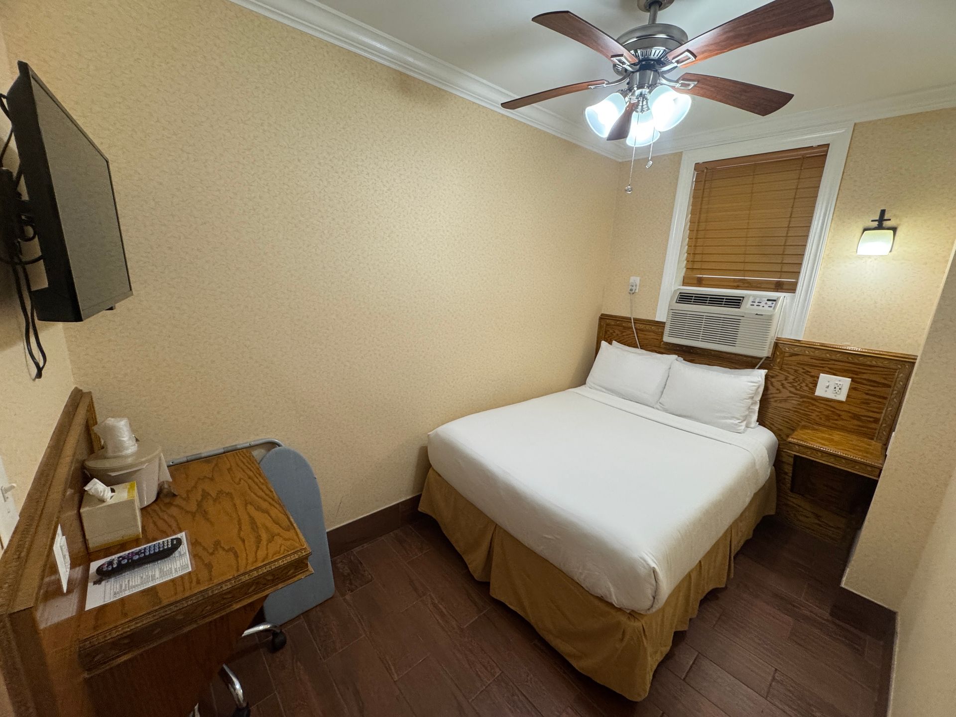 A small hotel room with a bed, desk, and mounted TV. The room has wooden floors and tan walls, with a window.
