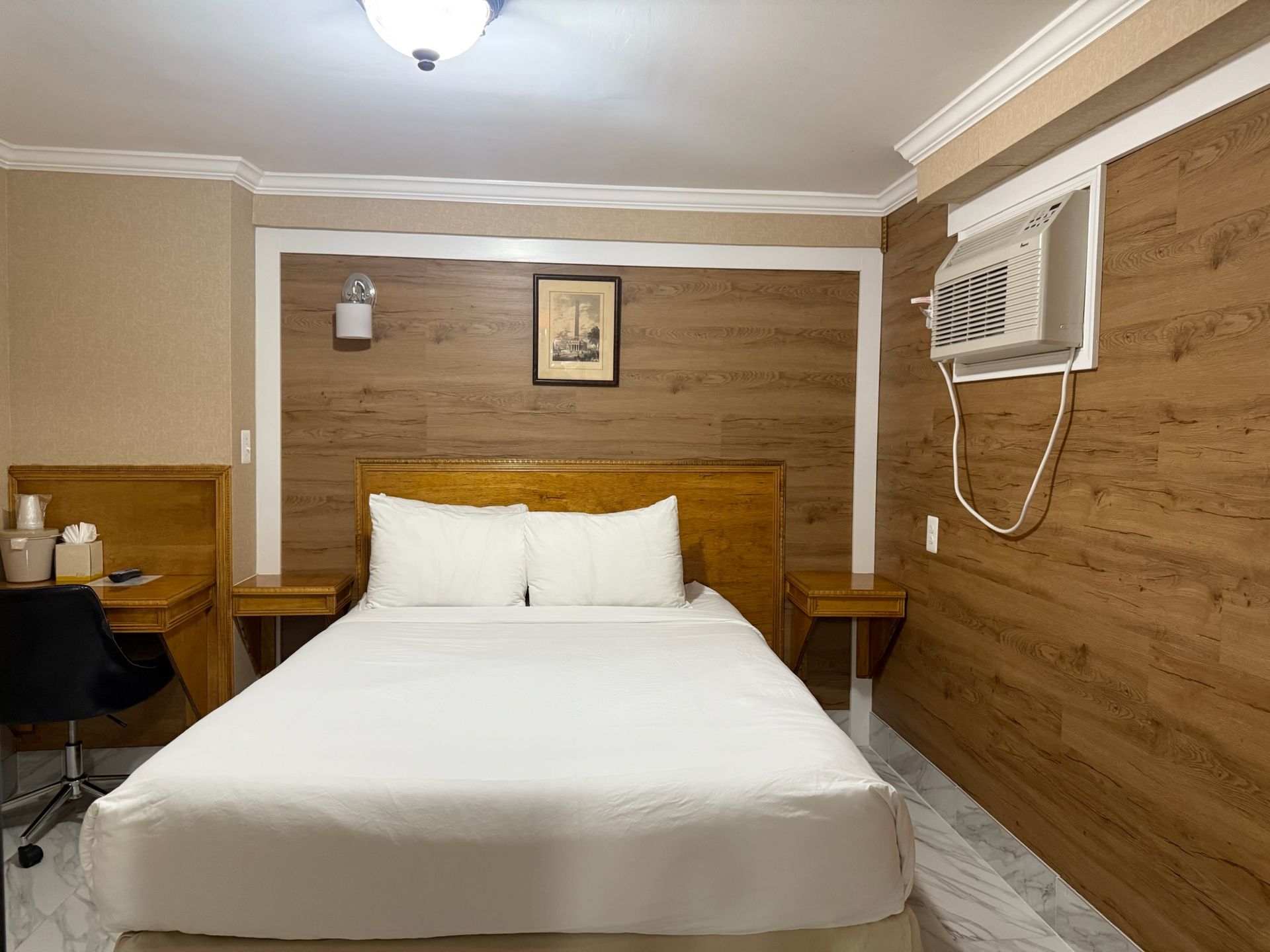 Hotel room with a bed, desk, and air conditioner. The walls have wood paneling and the bed has white linens.
