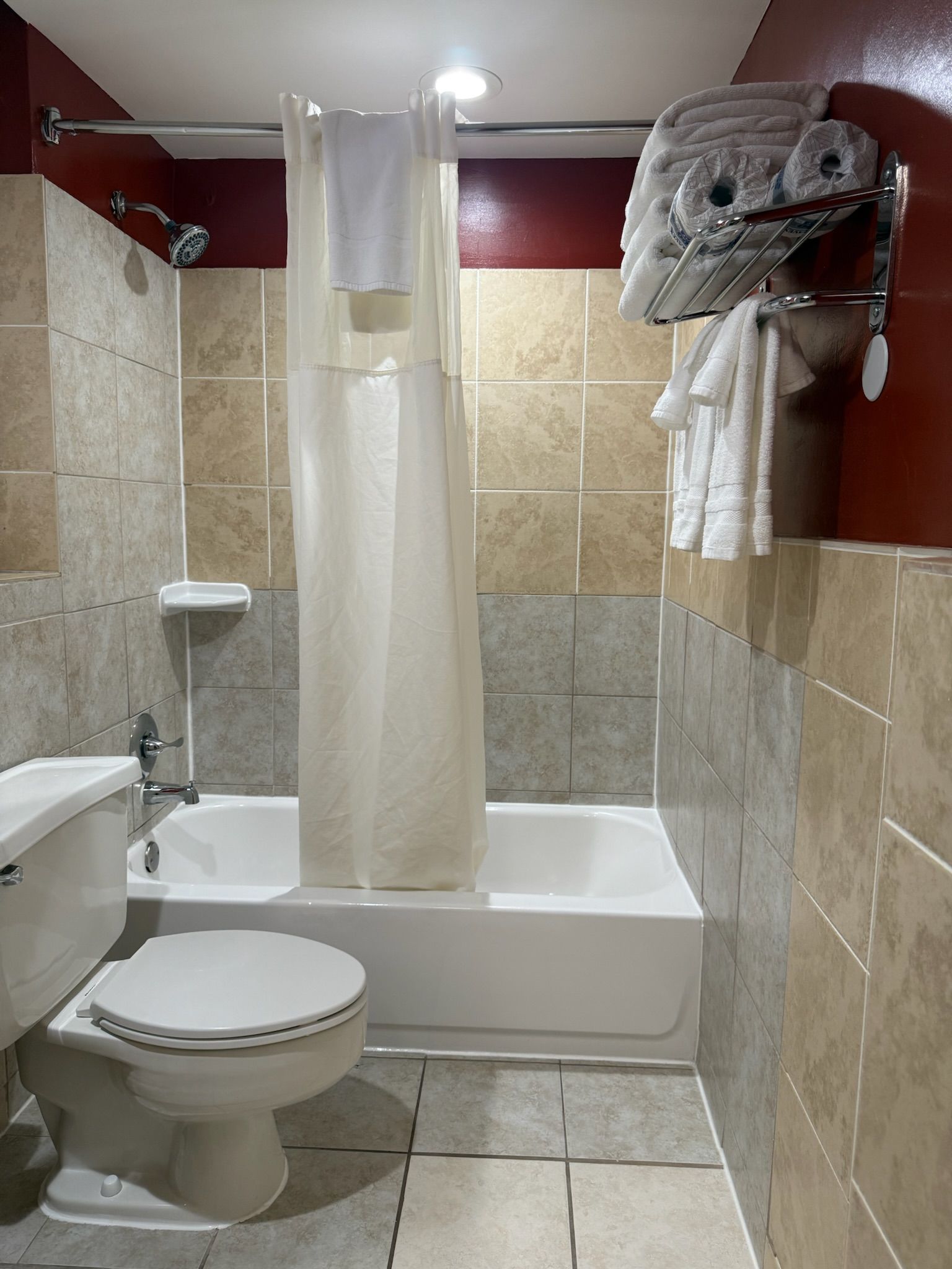 A small bathroom with a white bathtub, toilet, and beige tile walls. A shower curtain and towel rack with towels are visible.