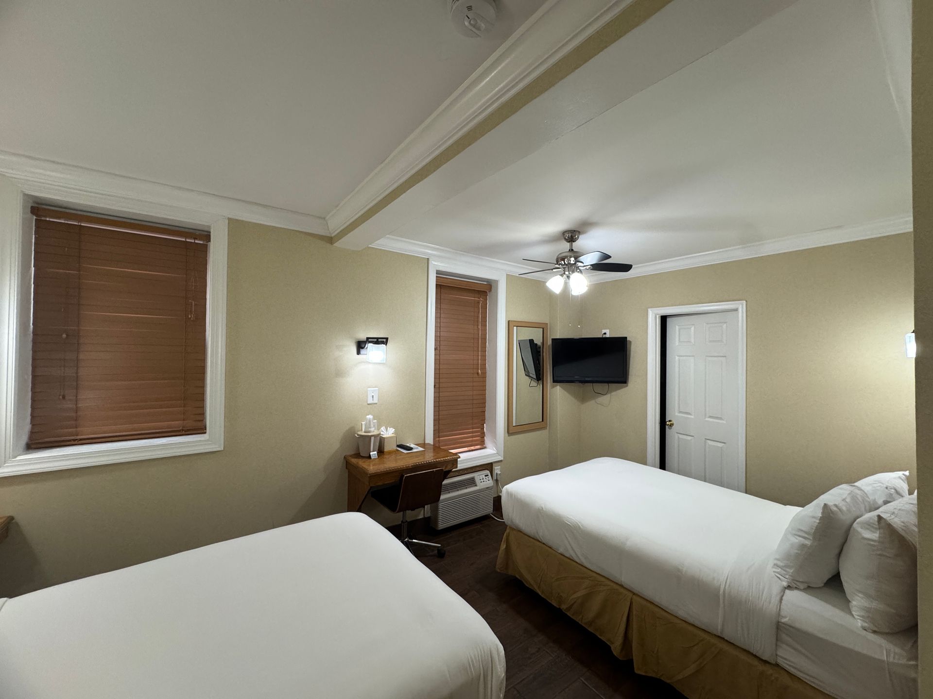 Hotel room with two twin beds, a desk, and a TV. The room has beige walls, brown blinds, and a ceiling fan.