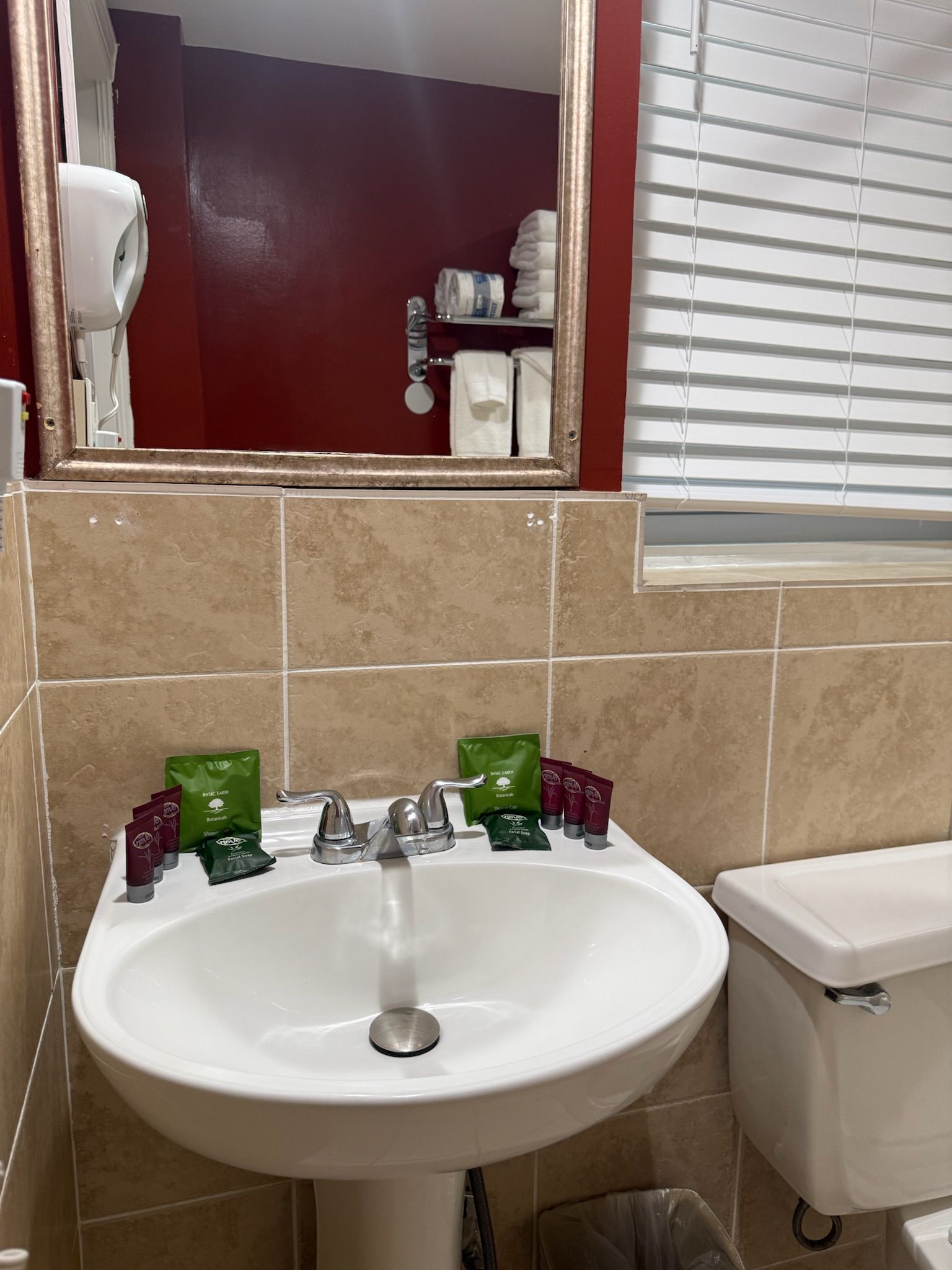 Bathroom sink with toiletries, mirror, and toilet. Beige tile, white sink and toilet, red wall reflected in the mirror.