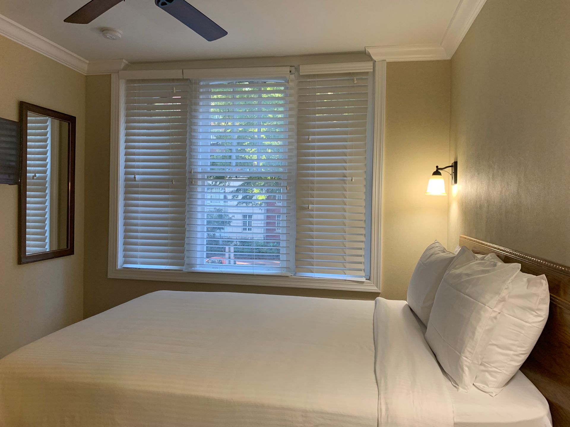 A hotel room with a bed, window with blinds, mirror, and a lamp. The walls are beige, and the bedding is white.