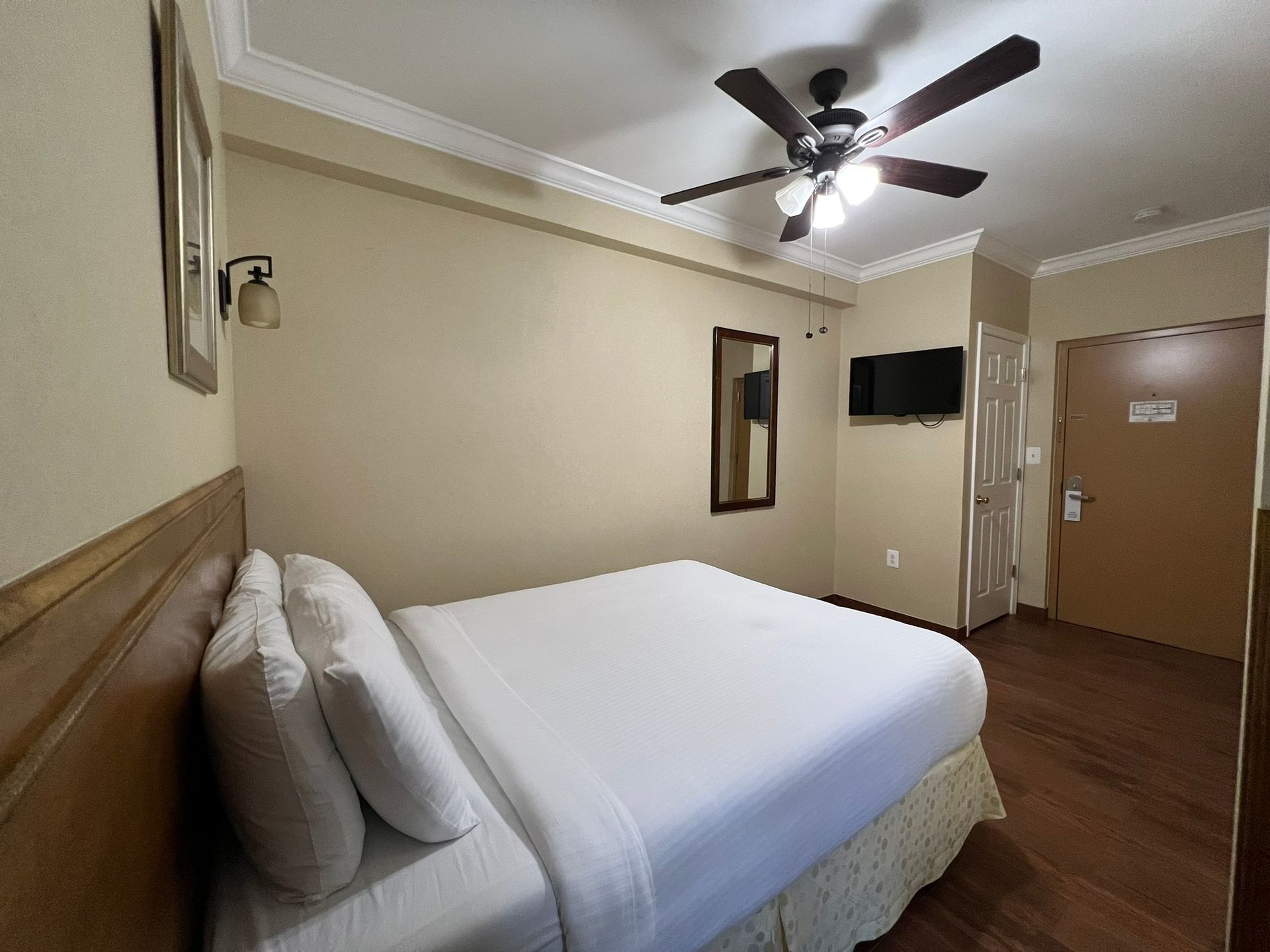 A hotel room with a bed, TV, ceiling fan, mirror, and closed door; neutral color scheme.