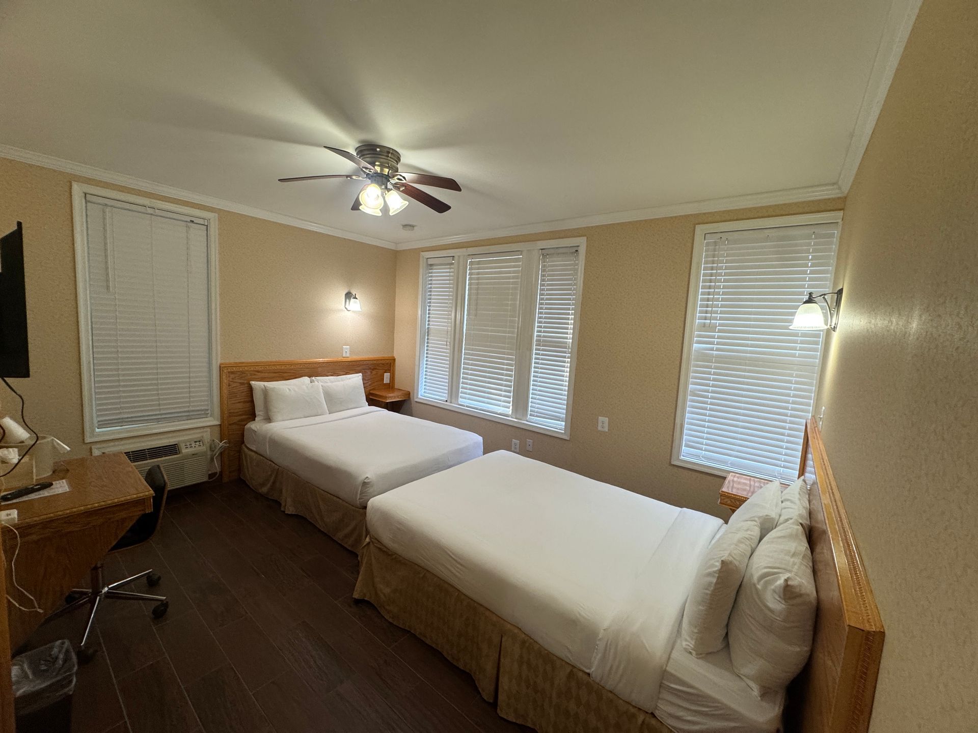 A hotel room with two beds, brown walls, and wood floors. White bedding and blinds, a ceiling fan, and sconces on the walls.