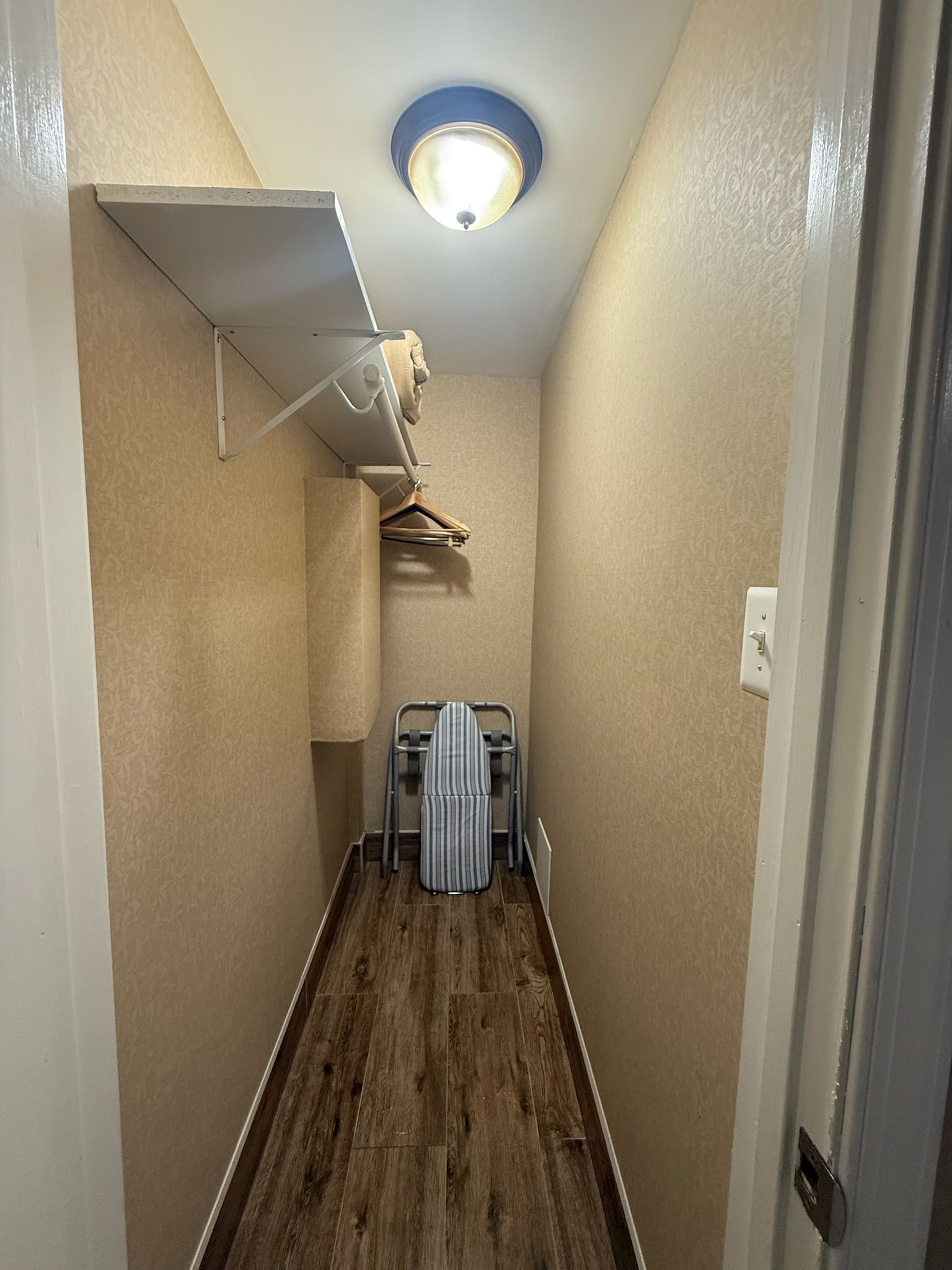 Narrow hallway with wood-look flooring and beige walls, a light fixture, shelf, ironing board, and hanging clothes.