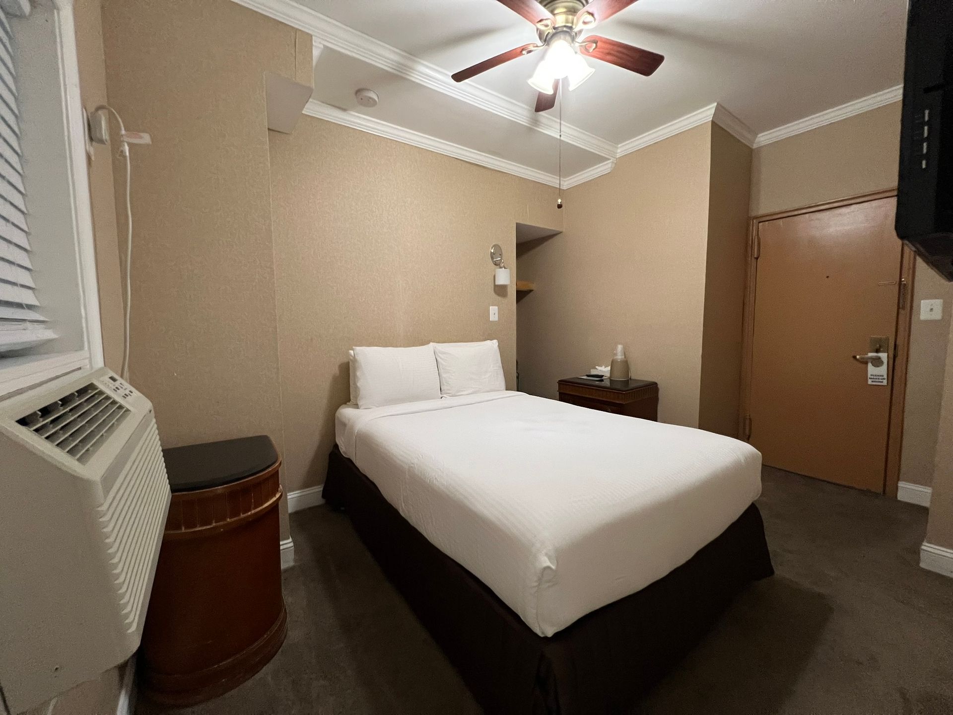 A small hotel room with a double bed, a window with blinds, and a brown door. Beige walls and ceiling with a bed skirt.
