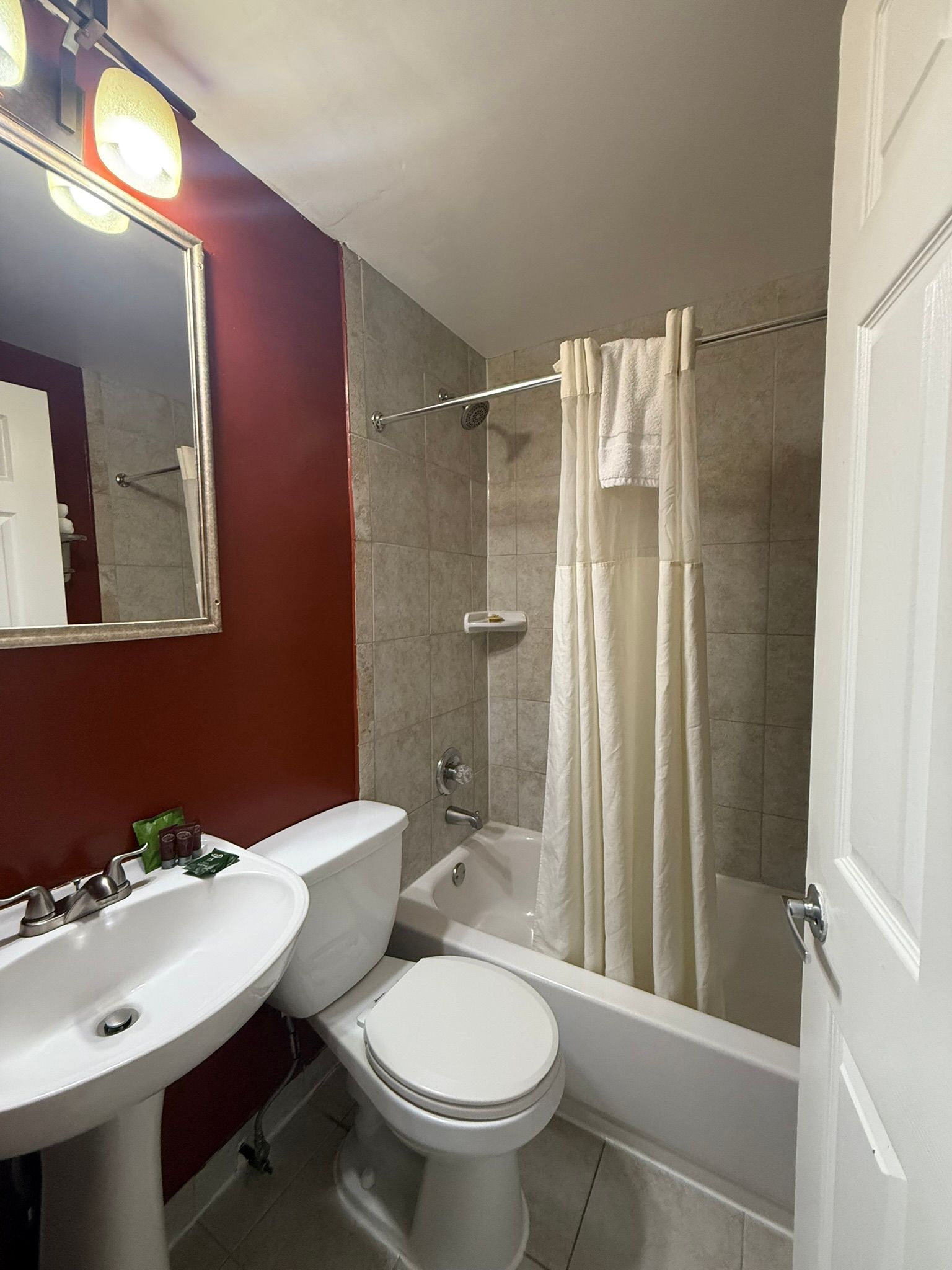 A small bathroom with a red wall, toilet, sink, and a bathtub with a shower curtain.