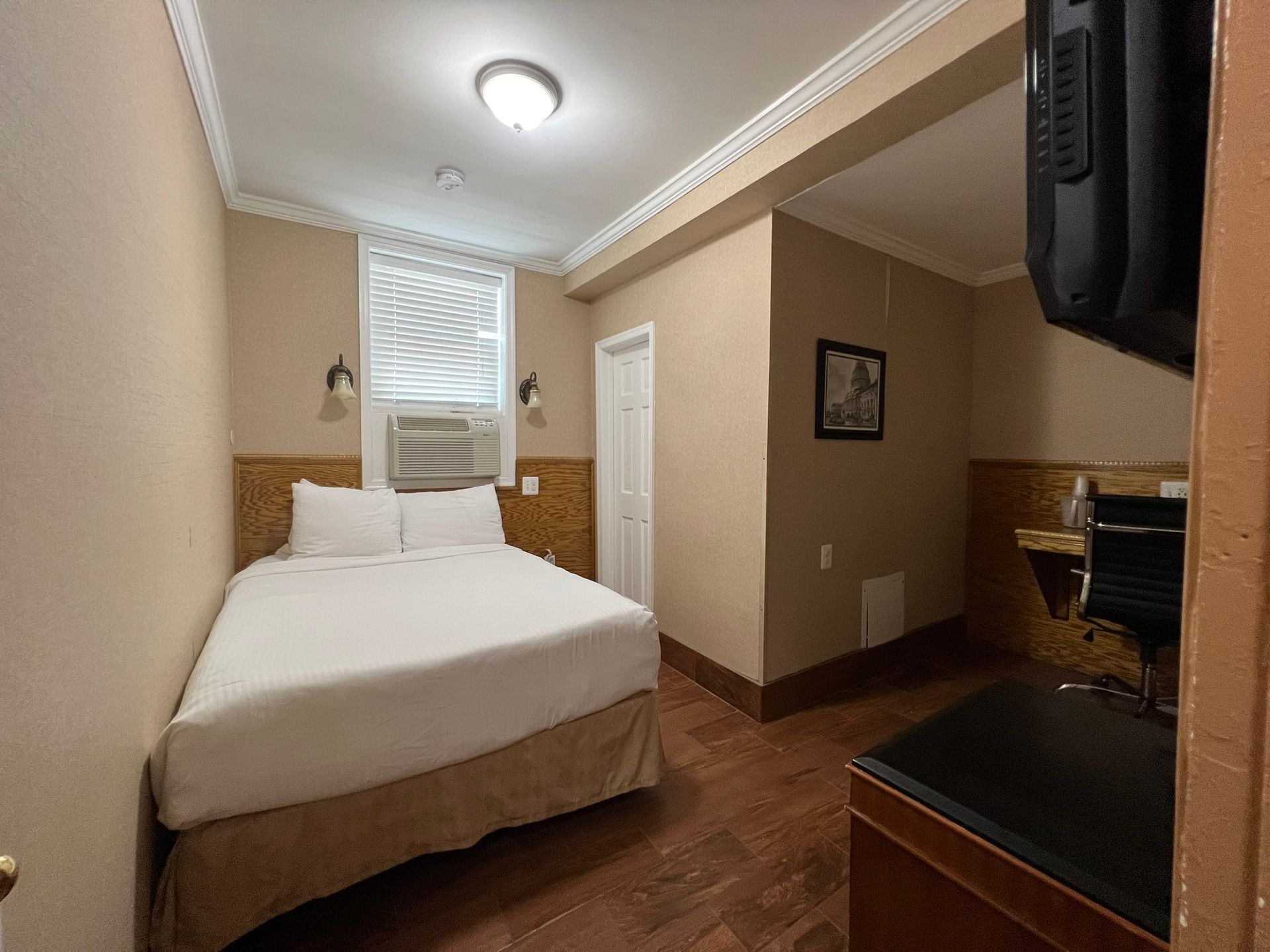 Small hotel room with a bed, desk, and wall-mounted TV. Beige walls, brown wood floor, and a window.