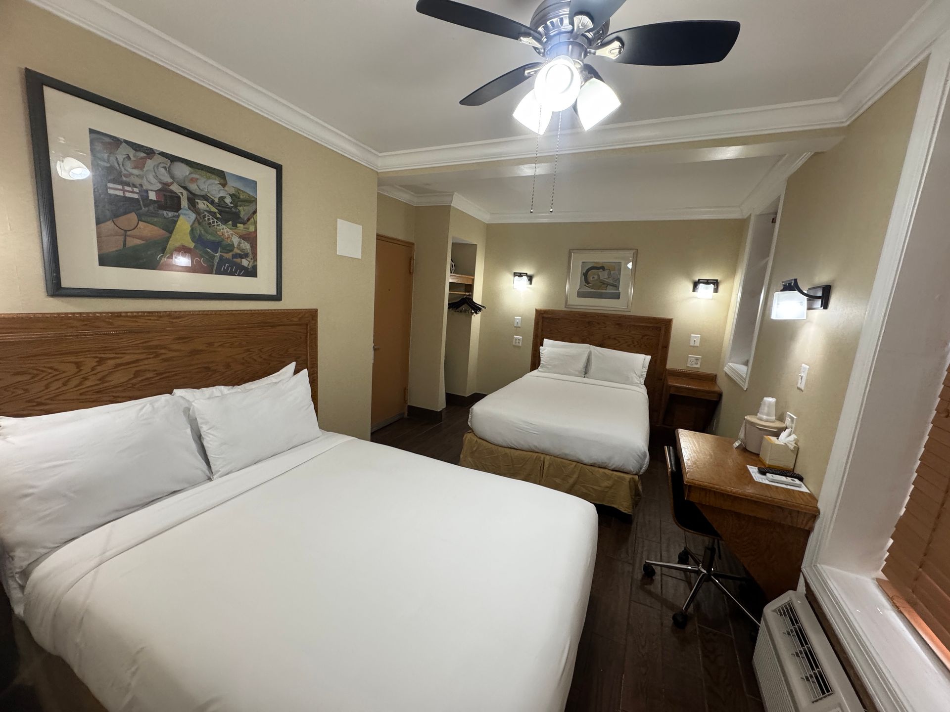 Hotel room with two beds, a desk, and a window. Walls are beige; furniture is wood-toned.